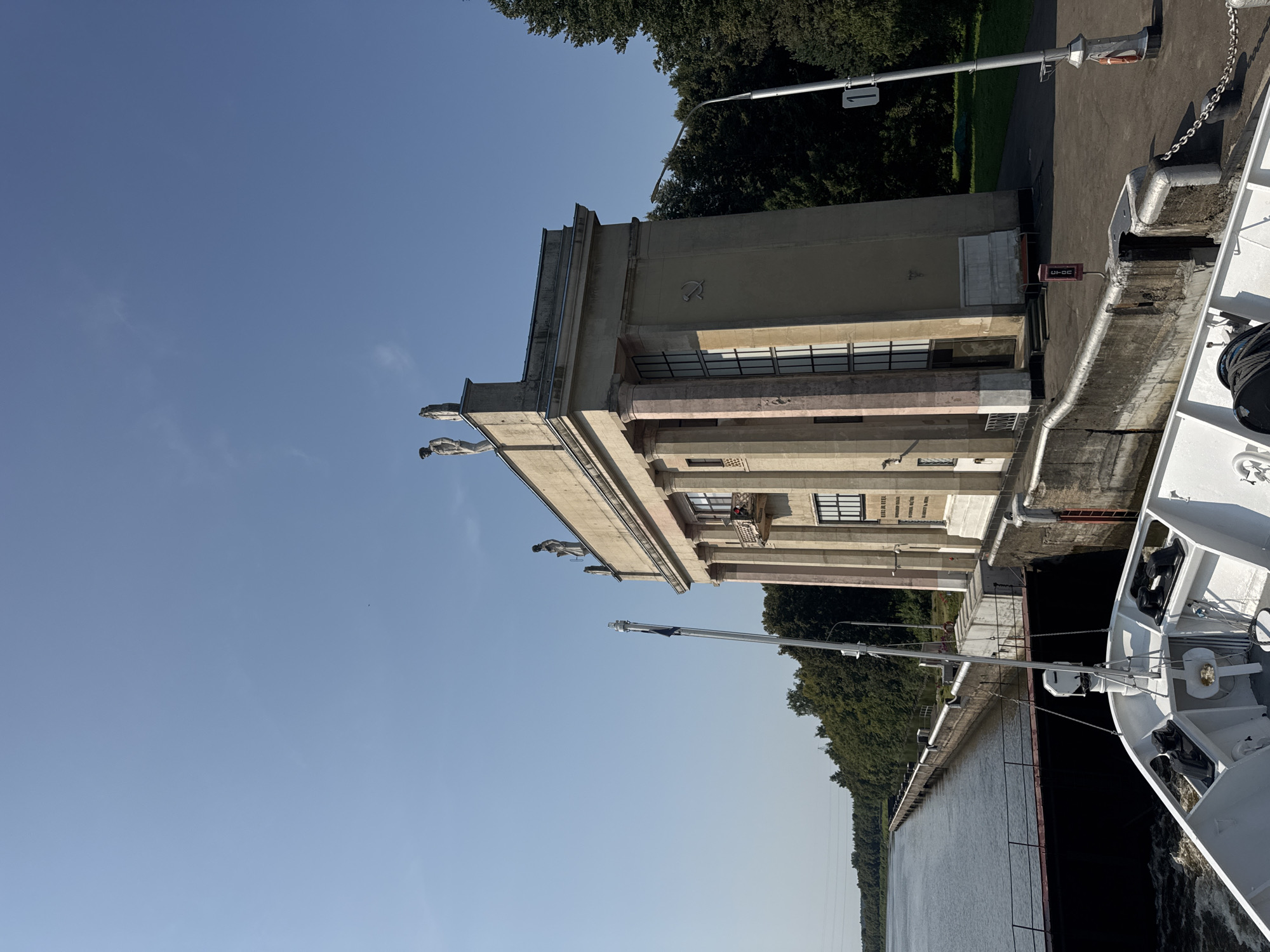 The image shows a perspective from a white boat entering a lock, likely at the Iksha Waterway Gate in Iksha, Russia. The lock gate is a large, imposing concrete structure with a classical facade, featuring tall columns and statues atop the roof. The building is painted a light beige color, with pinkish-tinged columns. A hammer and sickle symbol is visible on the facade. The water in the lock is choppy, indicating the boat is moving. Lush green trees line the banks of the canal in the background. The sky is a clear, bright blue. The scene is bathed in sunlight, suggesting a daytime setting.