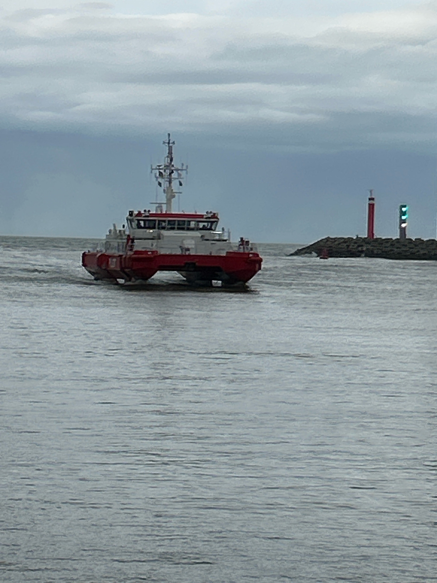 A modern, red and white catamaran vessel is navigating through choppy gray water. The boat has a multi-level cabin structure, with a prominent bridge section. In the background, a rocky jetty or breakwater is visible, topped with two navigation lights: a tall red lighthouse and a shorter green light array. The sky is overcast with heavy, gray clouds. The scene appears to be at the entrance to the port of Ostend, Belgium, given the presence of the navigational aids characteristic of such a location.
