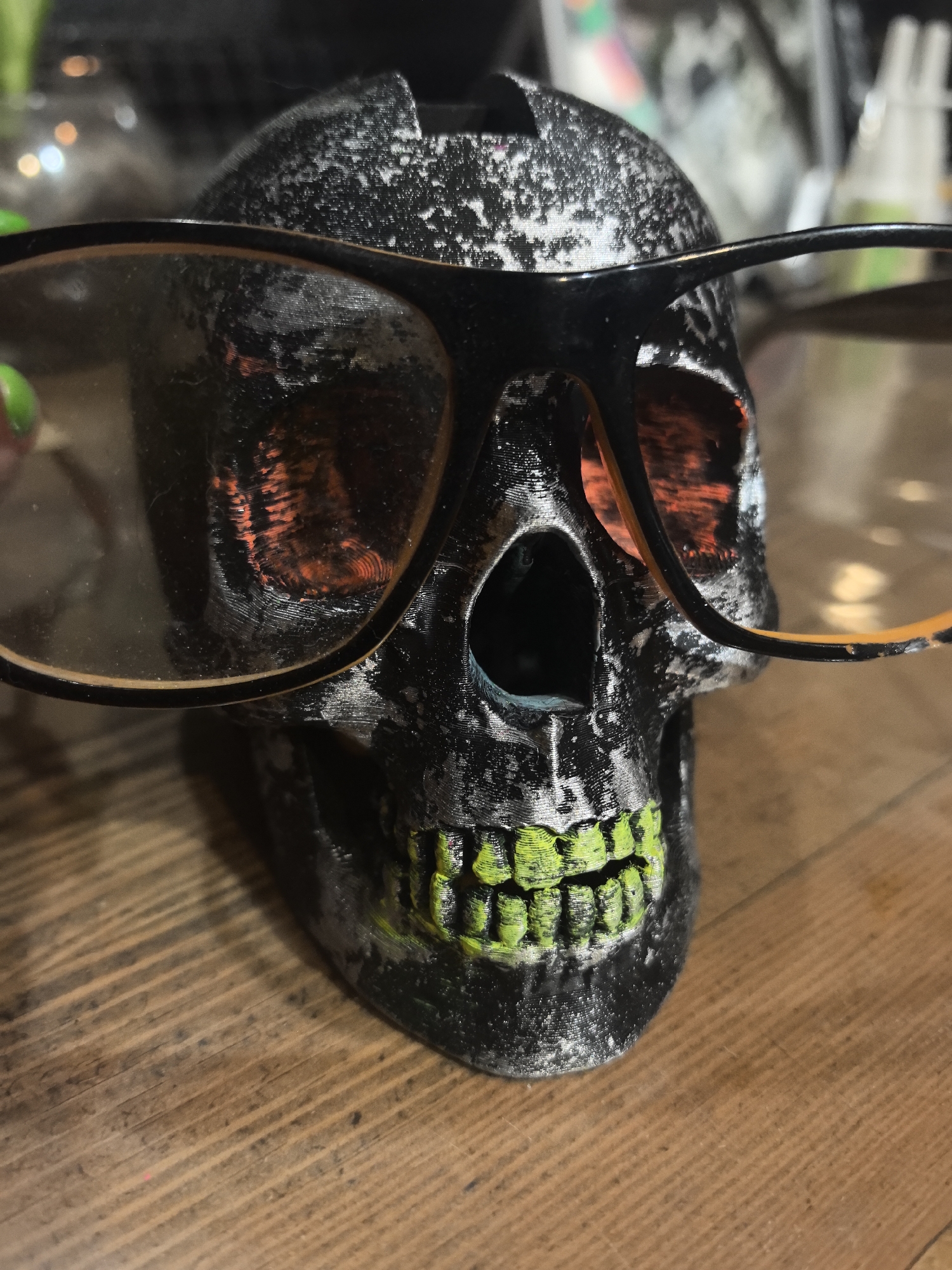 A close-up shot of a black and white skull with bright green teeth wearing black-rimmed glasses, resting on a wooden surface. The background is blurred and indistinct, but appears to show other objects. The image appears to be taken indoors. The city and country of Noviy, Russia are not evident in the image.