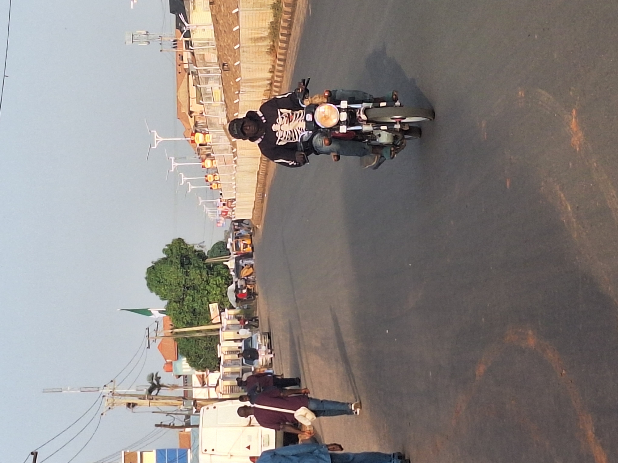 A daytime scene on a paved road in Sabon Tasha, Nigeria. A man wearing a black jacket with a white skeleton design rides a motorcycle towards the camera. Alongside the road, pedestrians walk and vehicles are parked. A Nigerian flag flies from a pole near trees and buildings. Streetlights line the road.