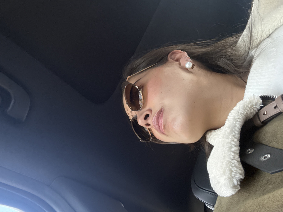 A low-angle shot of a woman wearing sunglasses and a white pearl earring. She is positioned in the lower-right corner of the frame with a dark gray ceiling in the background. She wears a light brown jacket with a white faux fur collar. A seatbelt with black and silver hardware is visible. Her lips are slightly parted, and her skin has a dewy finish. The sunglasses are round with gold frames and brown lenses.