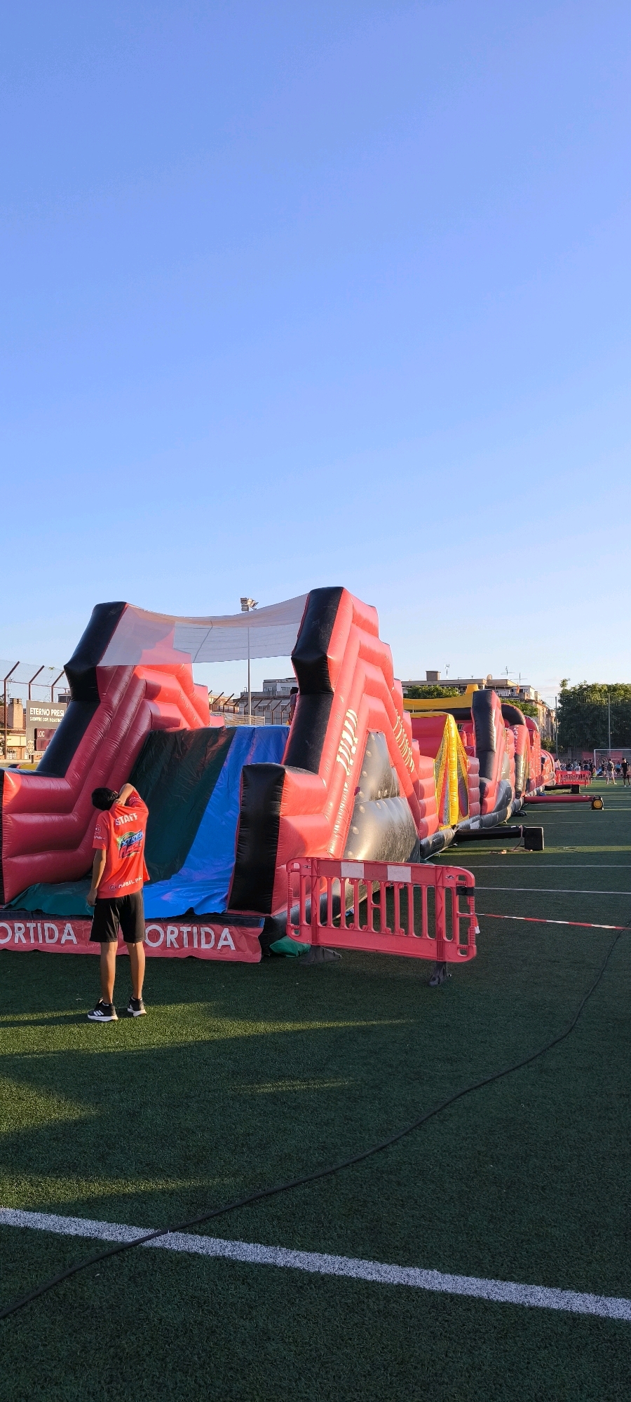 A person wearing a red shirt with "STAFF" written on it and black shorts stands on an artificial turf field, facing away from the camera and looking towards a large inflatable obstacle course. The obstacle course is red and black, with sections of blue and yellow. A pink plastic barrier fence is positioned in front of the inflatable structure. In the background, buildings and trees are visible under a clear blue sky. The setting appears to be an outdoor recreational area, likely a sports field, in Ripollet, Spain, given the context provided.