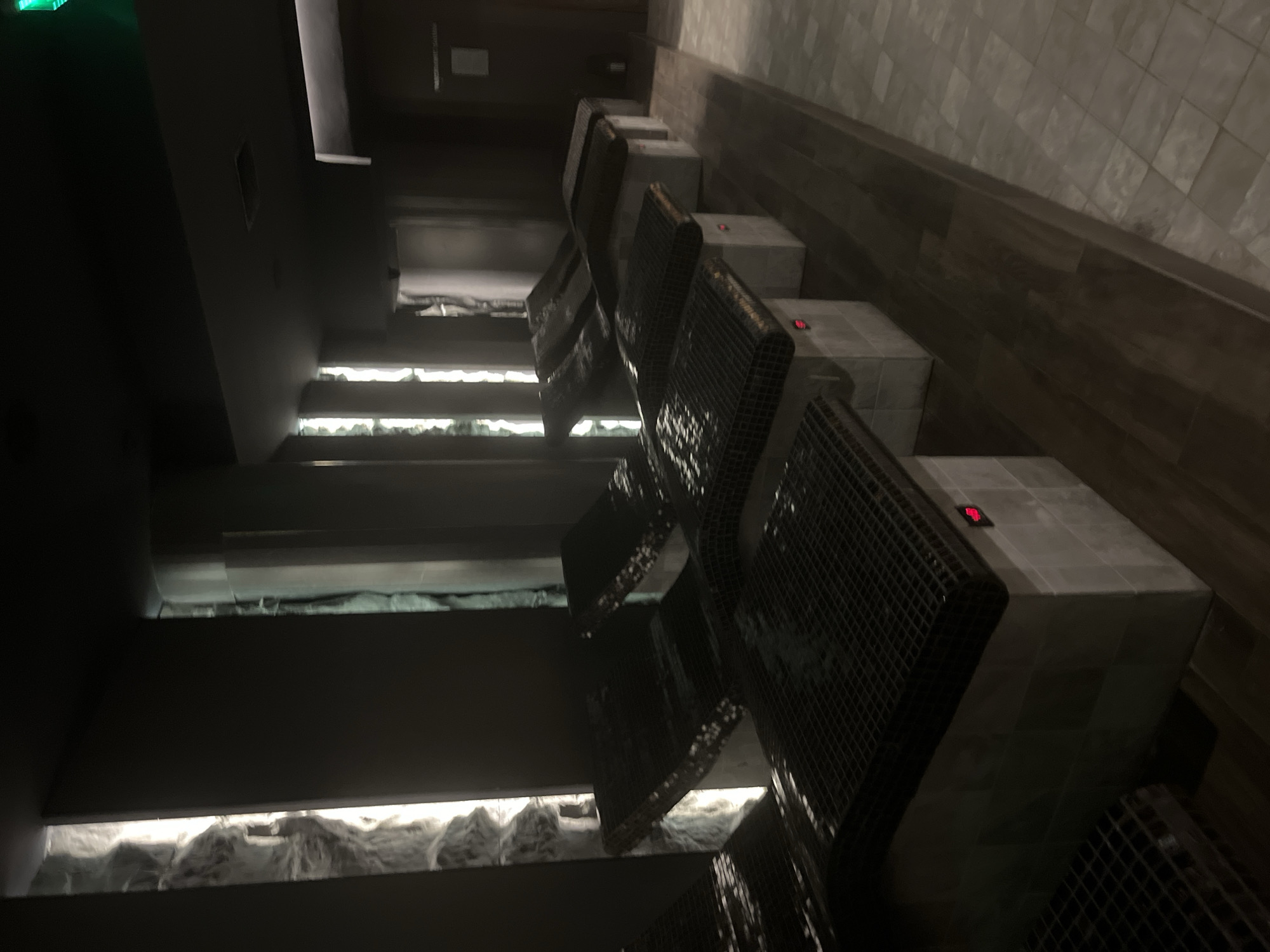 The image shows a sauna room. The room has tiered seating with tiled blocks and metal-mesh backrests. There is backlighting on the walls behind the seating, possibly with decorative rock or snow-like material. The walls are dark, and there is a green exit sign visible in the upper left corner. The tile on the wall looks like it could be in the city of Sofia, Bulgaria.