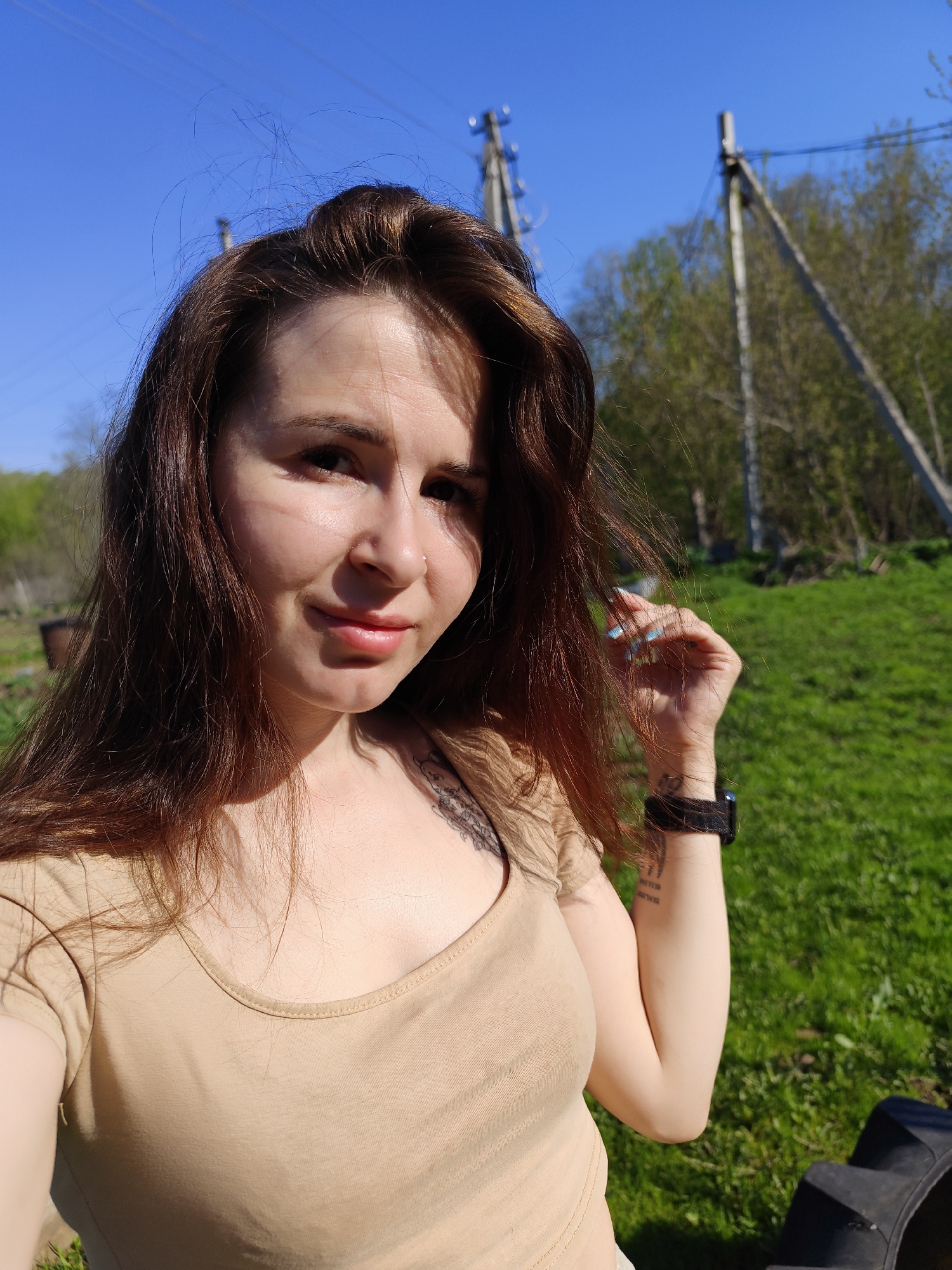 A young woman with long brown hair poses in a grassy, outdoor environment under a bright blue sky. She is wearing a tan-colored t-shirt and a black smartwatch on her left wrist. A tattoo is visible on her upper chest and arm. The background includes trees and wooden utility poles with electrical lines. The location is Фоки, Russia.