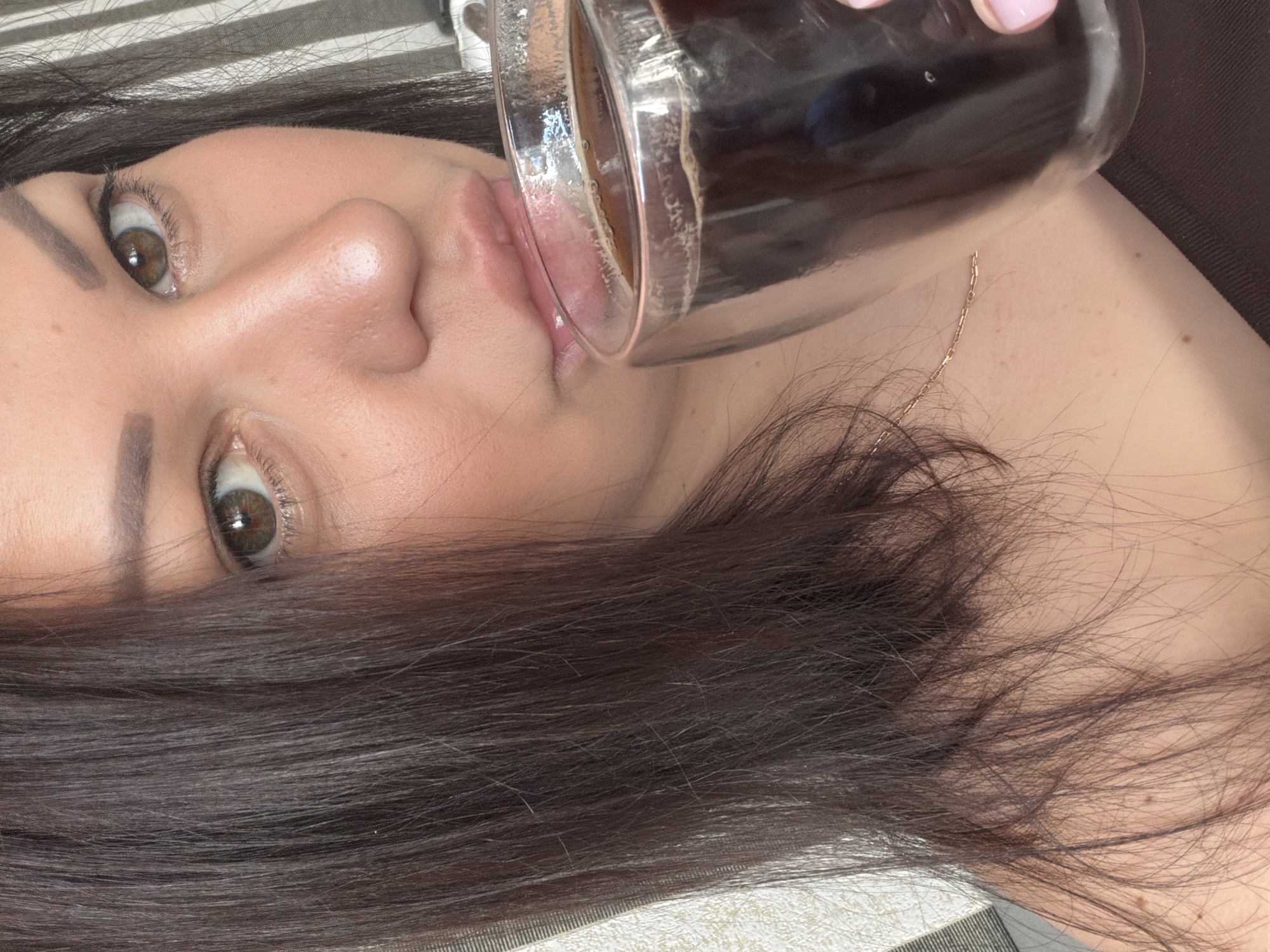 The scene is located in the city of Sochi, Russia. A woman with long, dark brown hair and hazel eyes is depicted in a close-up, chest-up shot. Her hair partially obscures the left side of her face. She has visible dark eyebrows and eyelashes. Her lips are slightly parted, in contact with the rim of a clear, double-walled glass mug. The mug contains a dark liquid, consistent with coffee, with some visible foam or bubbles on the surface. Her right hand, with light pink nail polish on the visible finger, holds the mug. She wears a delicate gold chain necklace around her neck and a dark, possibly brown, off-the-shoulder top. The background consists of a light-colored textured wall on the left and a vertical striped pattern on the right.