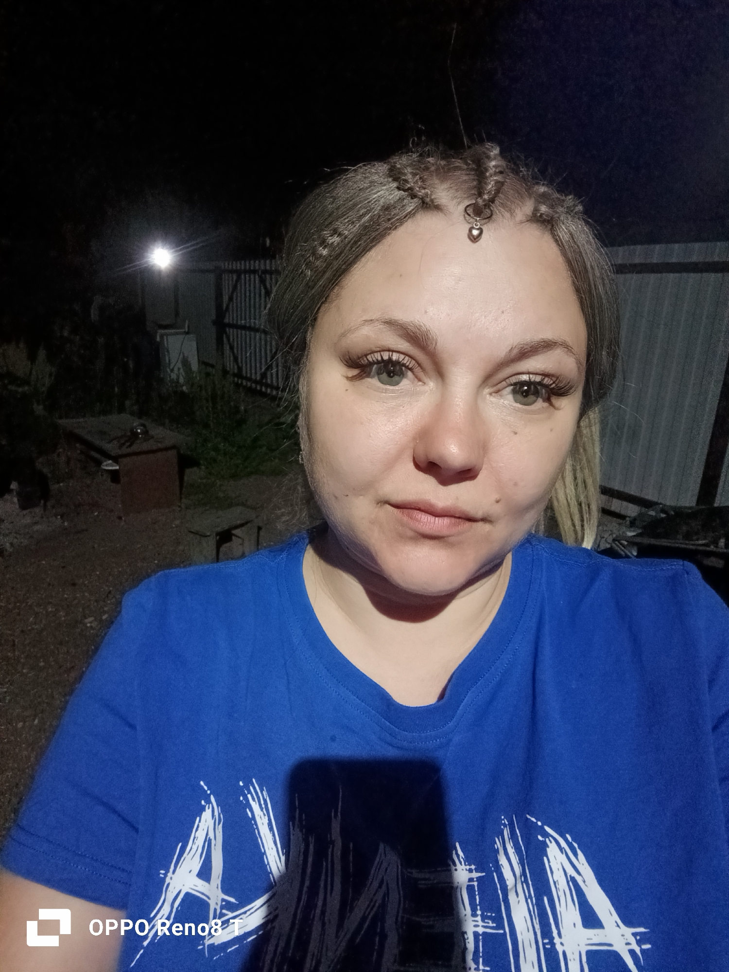 A woman with blonde hair styled in braids and a heart-shaped charm at her forehead, wearing a blue t-shirt, stands in an outdoor, possibly rural or industrial setting at night. The background includes a metal fence, some structures, and a bright light source. The image is a selfie, taken from the chest up, focusing on the woman's face and upper body.