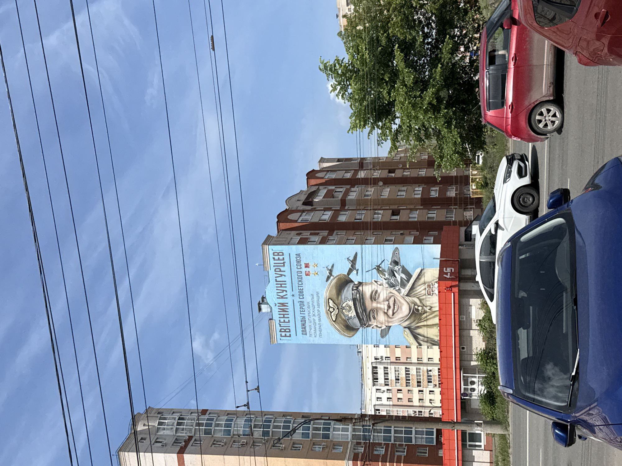 The image captures a street scene in Izhevsk, Russia, featuring a large mural of Evgeniy Kungurtsev, a decorated Soviet pilot and twice Hero of the Soviet Union, painted on the side of a multi-story building. The mural is positioned prominently against a blue sky background and includes images of aircraft associated with Kungurtsev's service. The foreground is filled with parked cars, including a blue car in the right corner, a white car partially visible, and a red car further in the background. Power lines stretch across the top left of the frame, adding a common urban element. The surrounding buildings are typical residential structures. The overall scene provides a glimpse into the local environment, where Soviet-era military figures are commemorated in public art.