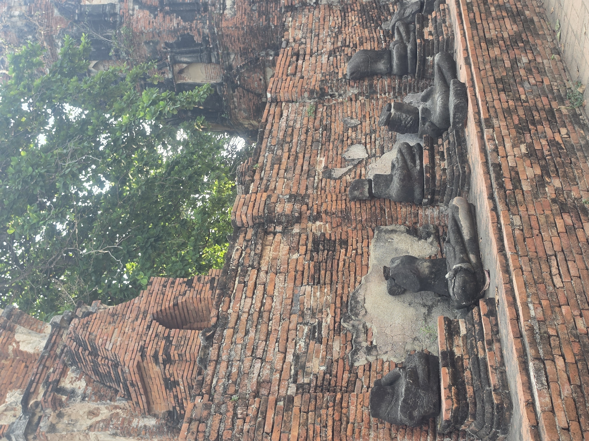 The image depicts the ruins of Wat Yai Chaimongkol in Phra Nakhon Si Ayutthaya City Municipality, Thailand. The ruins are constructed of red brick. The main elements are the brick walls and the remains of seated Buddha statues. The lower portion of the statues are visible. A tree with green leaves is also in the view, partially obscuring the brick structure. The sky is not visible. The image suggests a historical or archaeological site.