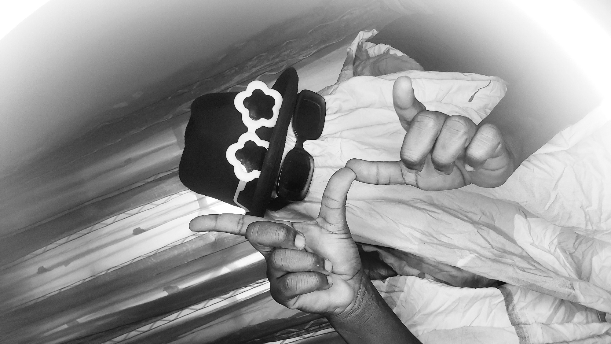 A person wearing a hat and novelty glasses, with their face obscured by fabric, is shown in a black and white, dimly lit indoor setting. The person's hands are positioned in the foreground, with fingers extended and making shapes. The background consists of draped fabric, possibly curtains. The image is a close-up, with some areas blurred by light. There is no indication of the location being in Narayi, Nigeria.