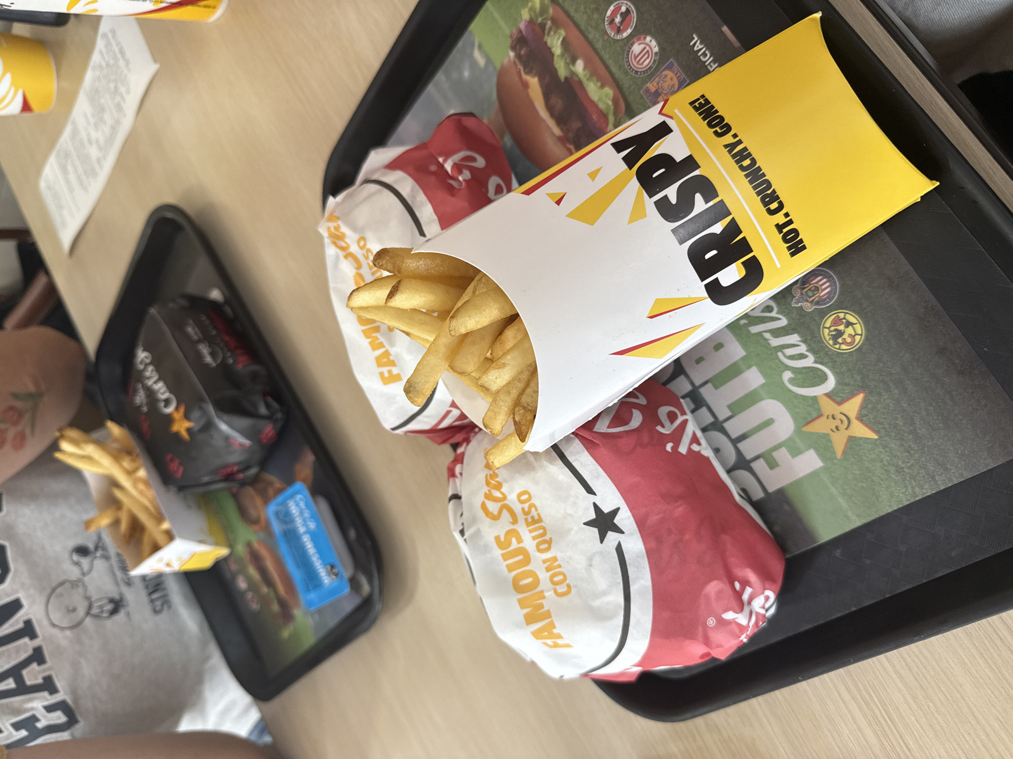 The image shows a meal from Carl's Jr. on a black tray. The meal includes a box of french fries with "CRISPY" printed on it, and two Famous Star with Cheese burgers. A smaller tray also has a burger and fries. The photo was likely taken at a Carl's Jr. in San Bernardino Tlaxcalancingo, Mexico.