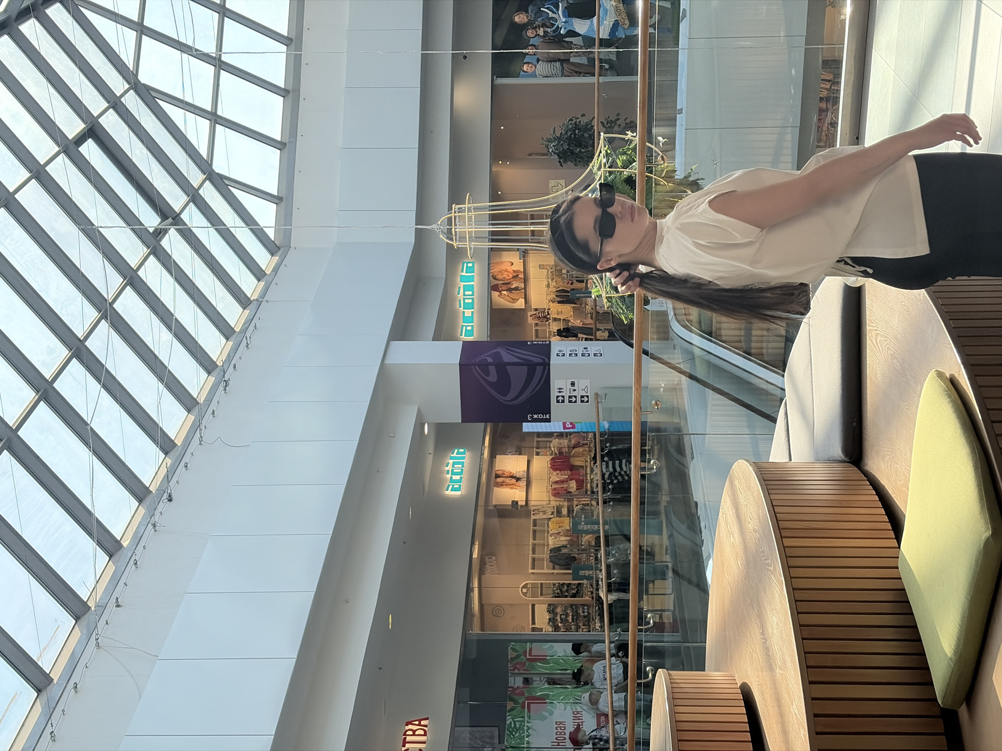 The image depicts an indoor scene in Izhevsk, Russia. A young woman with long brown hair, sunglasses, a white t-shirt, and black pants is sitting on a wooden bench with a light green cushion. She is looking at the camera. The bench is part of a round seating area with wooden slats. Behind her is a glass barrier. Through the glass, there is a multi-level shopping mall interior. Signage for shops like "Ostin" is visible. The mall has a high, sloped ceiling with a grid of glass panels. There is a decorative hanging light fixture.