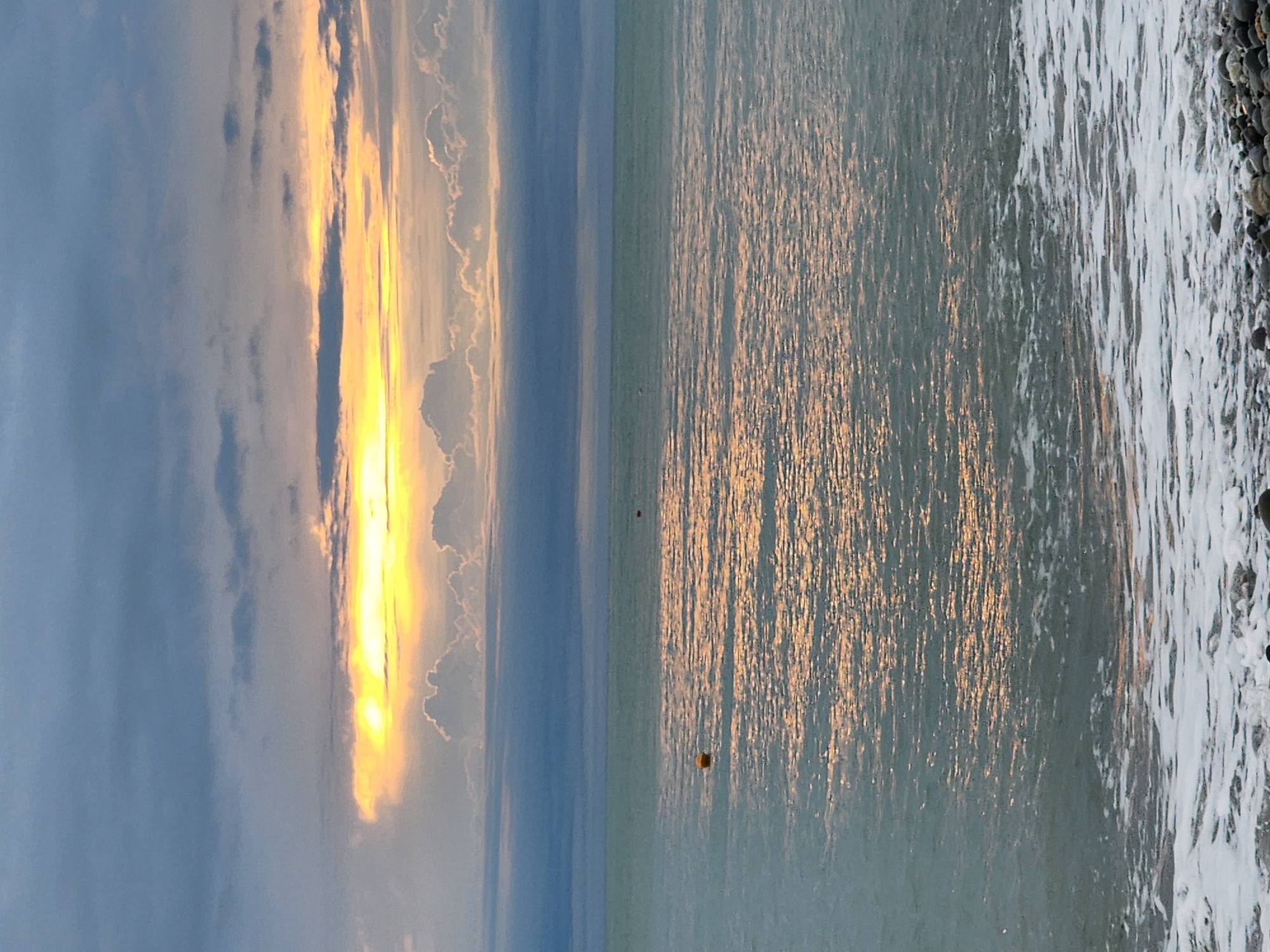 This image depicts a serene coastal landscape at either sunrise or sunset in Sochi, Russia. The scene is dominated by the interplay of light on the sky and sea, with a pebble beach in the foreground.
In the upper portion of the frame, the sky features a dramatic display of light. A strong, golden glow, presumably from the sun, pierces through a band of clouds, creating a vibrant vertical streak of illumination. This bright light contrasts with the softer blue and grey hues of the surrounding clouds and sky.
Below the horizon line, which stretches horizontally across the middle, lies the sea. It reflects the golden sunlight beautifully, forming a shimmering, warm path across the water towards the viewer. The water appears relatively calm, with gentle ripples visible from the sun's reflection. Two buoys are present in the water: a distinct yellow, square-shaped buoy positioned closer to the viewer, and a smaller, dark, possibly red or orange buoy further out towards the horizon.
In the bottom right corner, a section of a pebble beach is visible. Foamy white waves are breaking and receding from the shore, revealing dark, wet stones. The texture of the frothy water contrasts with the smooth, rounded pebbles.
The time of day is clearly indicated by the golden hour lighting, suggesting a peaceful and majestic atmosphere. There are no people or visible text in the image.