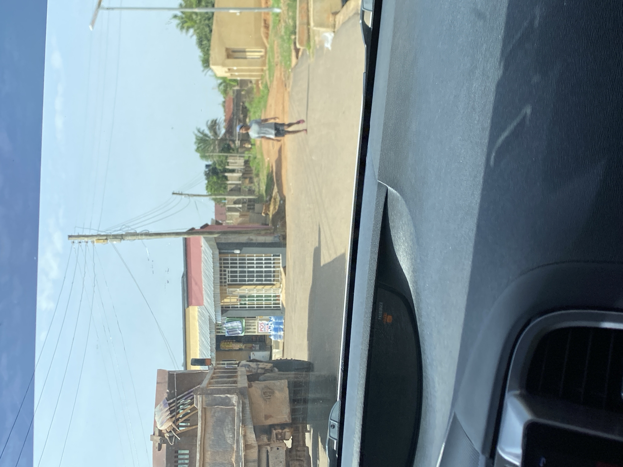 A person walks down a dusty road in Bar Jirgi Summa, Nigeria, casting a shadow on the asphalt. To the left, a dump truck is parked in front of a building with a metal awning. In the background, several buildings line the road, and utility poles with wires stretch across the sky. The scene is illuminated by bright sunlight, suggesting a clear day.