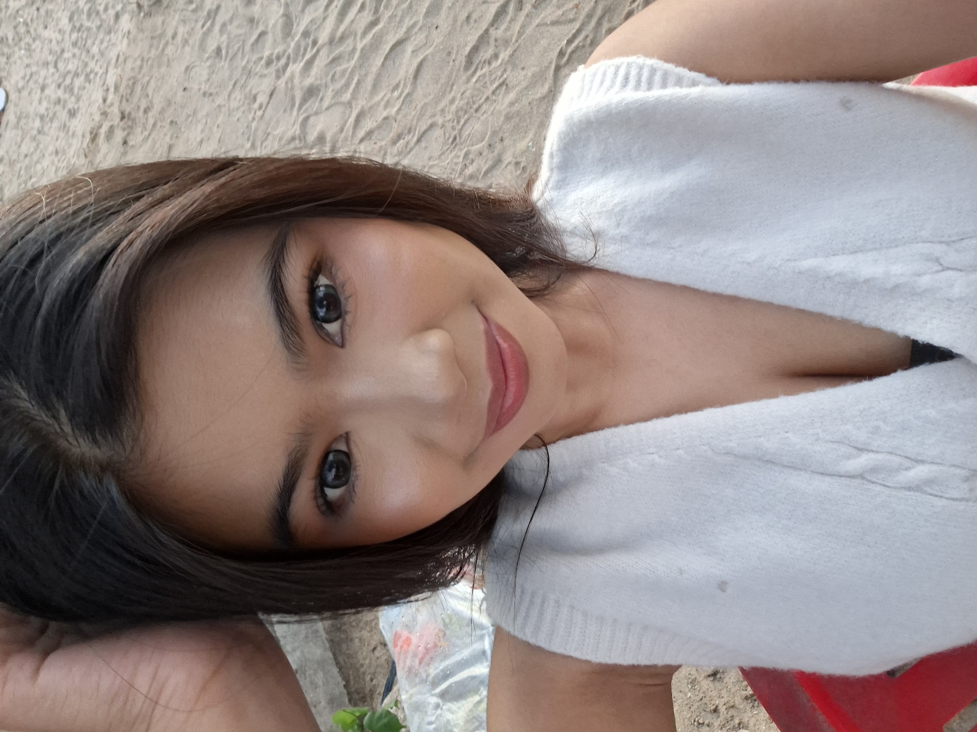 The image shows a close-up, selfie-style shot of a young woman with dark hair and makeup, wearing a white sleeveless top. She is positioned in the foreground, with her head and shoulders filling most of the frame. Her left hand is raised and resting on the top of her head. She is looking directly at the camera and smiling slightly. The background consists of sand, suggesting an outdoor setting. The lighting appears natural, possibly from daylight. The sand texture and the presence of what looks like a beach in the background are consistent with the environment of Kuta, Indonesia, a popular tourist destination known for its beaches.