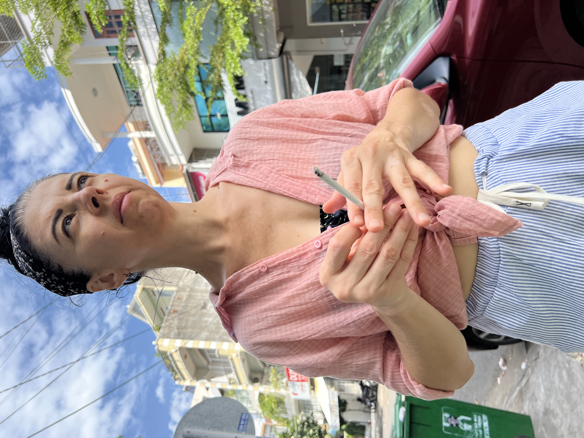 A woman with dark hair tied up, wearing a pink crinkled short-sleeved button-down shirt tied at the waist and blue and white striped loose-fitting pants, stands on a street in Nha Trang, Vietnam. She is holding a cigarette in her hands. The background shows buildings with multiple stories, some with balconies, and trees. A red car is parked behind her. A green trash bin is visible to her left. The sky is blue with scattered clouds.