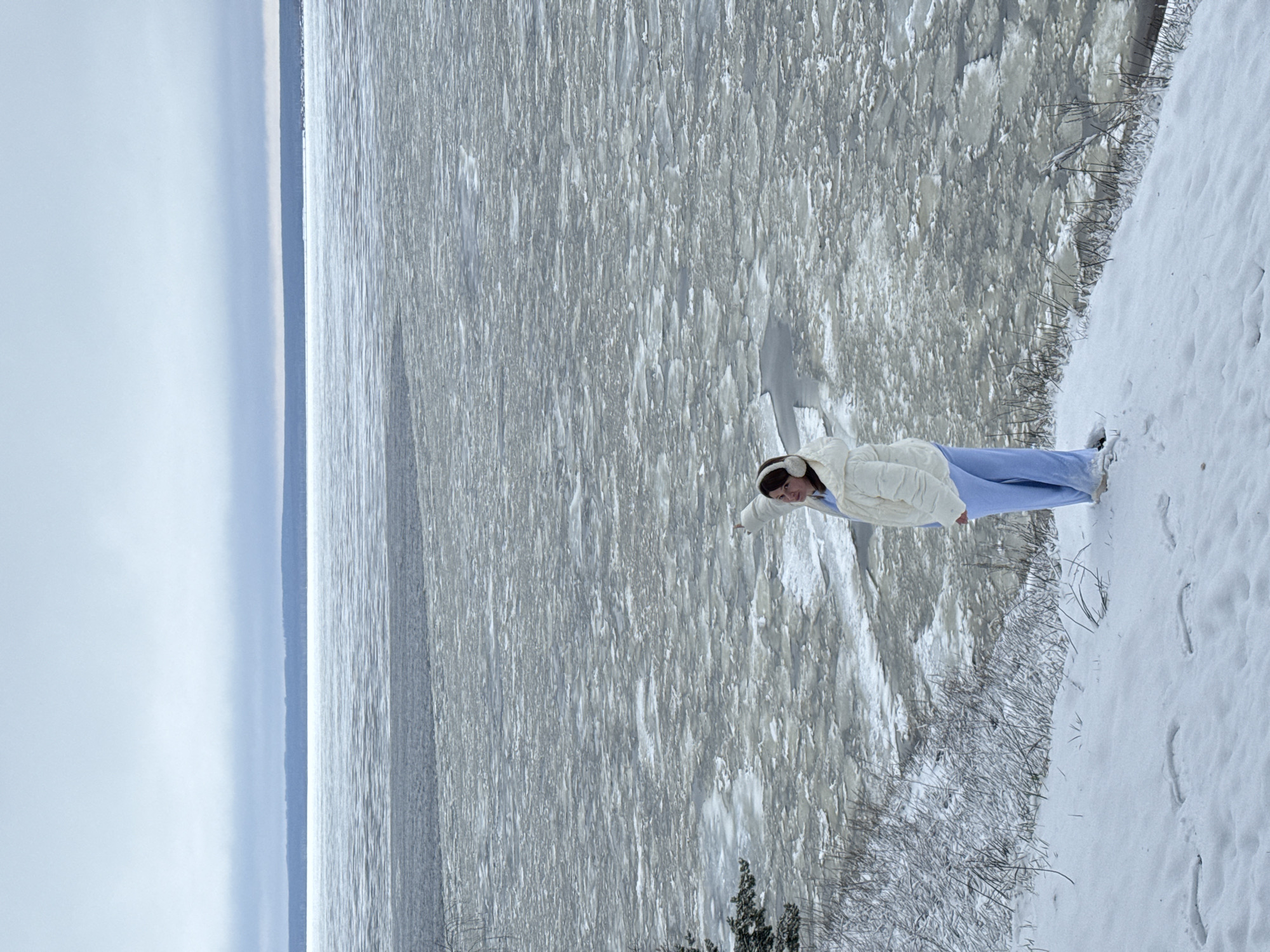 A medium, oblique shot shows a woman standing in the snow next to a body of water. The woman is wearing a white jacket, light blue pants, and white earmuffs. She is looking towards the water, and her right hand is pointed towards the water. The body of water appears to be covered in ice floes and stretches to the horizon under a gray sky. The snow-covered ground slopes downward towards the water, with bare branches and small trees visible. The location is Noviy, Russia.