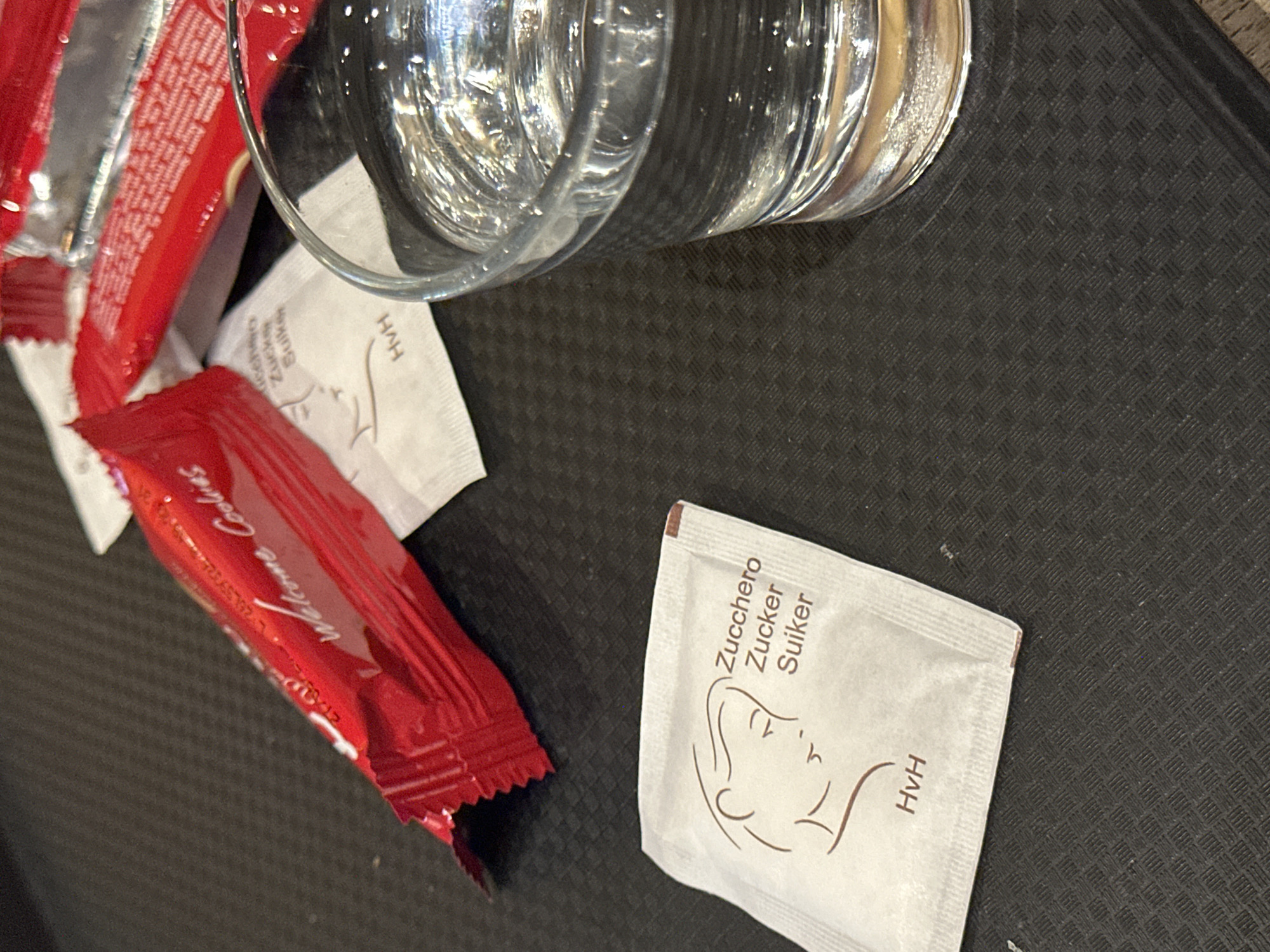 A close-up, eye-level shot shows a dark grey, textured placemat with several items on it. In the upper left, a partially opened red wrapper with the words "Welcome Cookies" in white script is visible. To the right of the cookie wrapper are two small, white packets, each with an imprint of a face and text that reads "Zucchero Zucker Suiker" and "HvH". Further to the right, a clear glass is filled with water, reflecting light. The overall scene suggests a refreshment or snack setting, possibly in Krefeld, Germany.