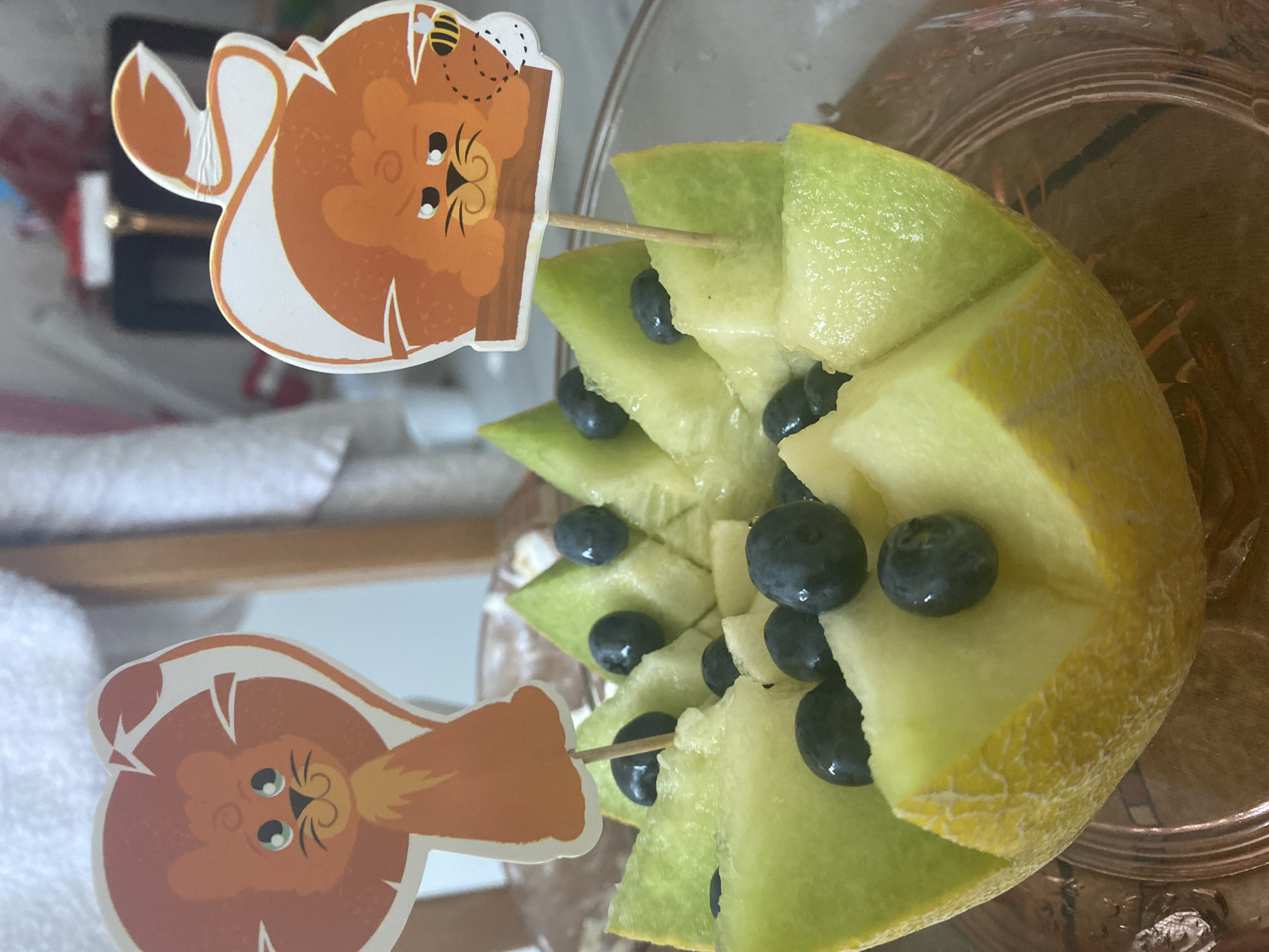 The image displays a close-up of a cantaloupe carved into a bowl-like shape, filled with pieces of melon and blueberries. Two decorative toothpicks with cartoon lion faces are inserted into the melon. The lion on the left has a friendly expression with big green eyes, while the lion on the right appears slightly grumpy and has a bee flying near its head. The cantaloupe is presented in a clear glass bowl, which sits on a wooden surface. The background is blurred but suggests an indoor setting with hints of furniture and possibly a window. The lighting is natural, coming from the front and slightly to the right. There are no discernible features that place this image specifically in Blagoevgrad, Bulgaria; it appears to be a generic indoor scene.