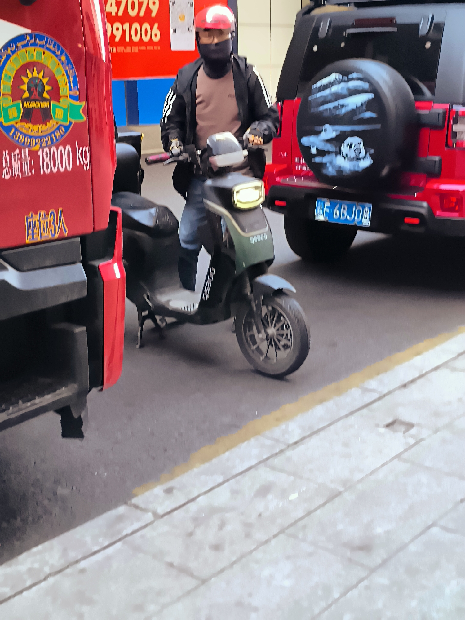 The image, taken in Ürümqi, China, shows a person riding a green electric scooter in the foreground, wearing a red helmet and a black face mask. The scooter has "G5000" written on its side. To the left, a red truck with Chinese characters and "total weight: 18000 kg" is partially visible, along with a circular logo. In the background, a red Jeep Wrangler is parked, with a spare tire cover decorated with a black and white image that appears to depict a landscape or a scene with a skull. The Jeep has a blue license plate that reads "MF 6BJ08". The scene takes place on a paved street with a yellow-lined pedestrian walkway.