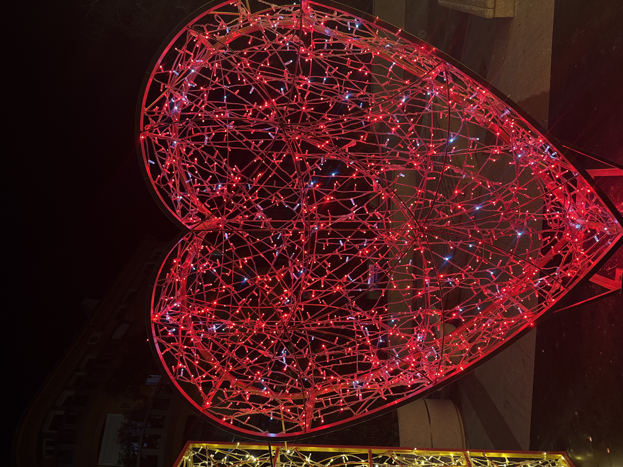 A large, heart-shaped decoration made of red lights is displayed outdoors at night. The heart is intricate, with many strands of lights crisscrossing to form its shape. Some white lights are also visible interspersed with the red. To the left, part of another decorative light structure, possibly a box shape, is visible, illuminated with yellow lights. In the background, a building with balconies and windows is dimly lit against a dark sky. The ground appears to be paved. This decoration is located at the Sea Garden in Burgas, Bulgaria, as part of the Christmas decorations.
