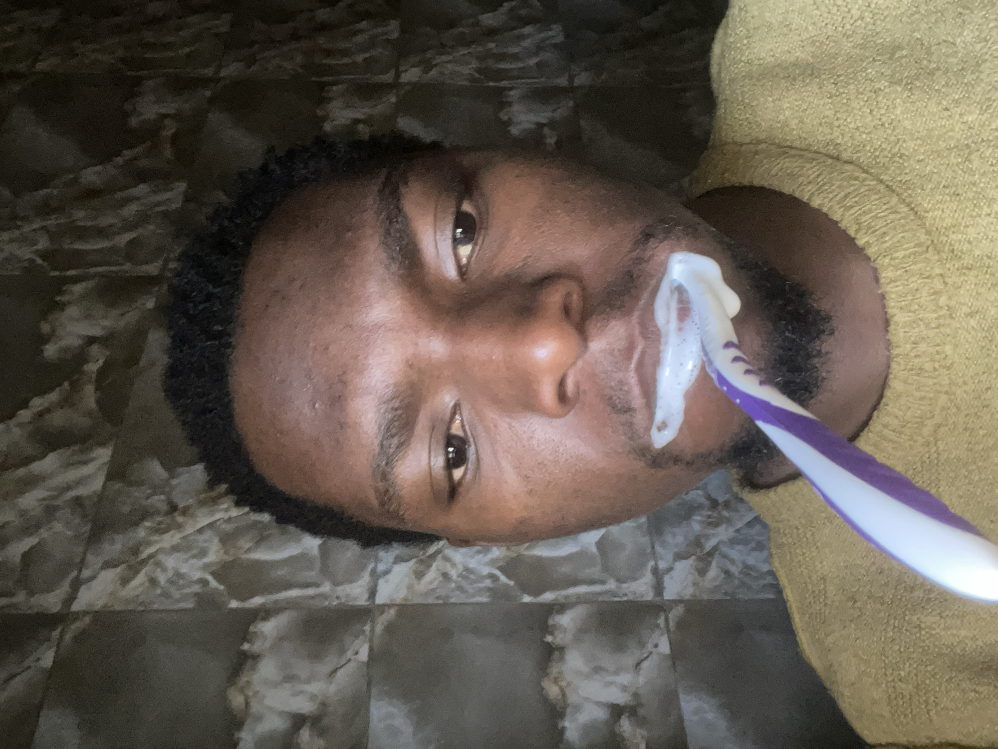 A close-up, rotated portrait shows a young Black man brushing his teeth in an indoor setting in Jos, Nigeria. He is positioned horizontally in the frame, with his head tilted slightly. He has dark, short, curly hair and a light beard/stubble, with dark brown skin. He wears a mustard yellow or light brown textured sweater, visible on his right side.
A white and purple toothbrush is held in his mouth, with white toothpaste foam visible around his lips and on the brush bristles. His eyes are open, looking directly towards the camera, and his expression is neutral, focused on the activity.
The background features dark, mottled, patterned tiles, suggesting a floor or wall in an indoor environment. The lighting is dim, contributing to a somewhat subdued atmosphere. No other individuals or objects are visible, and there is no visible text. The image captures a personal moment of daily hygiene.