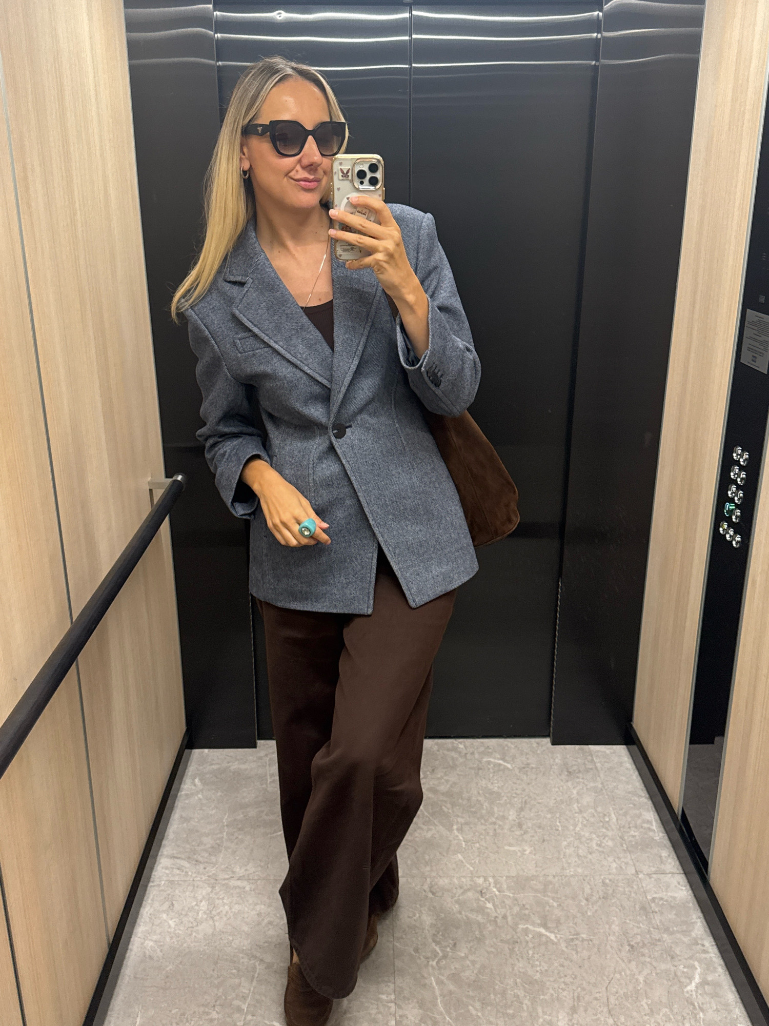 In an elevator, a woman with blonde hair and sunglasses takes a selfie. She is wearing a gray blazer over a brown top and brown pants, with a brown bag slung over her shoulder. She is also wearing a large turquoise ring on her right hand. The elevator walls are a mix of dark gray and light wood paneling, and the floor is tiled in a light gray marble pattern. The elevator controls are visible on the right side of the frame, featuring a series of illuminated buttons.