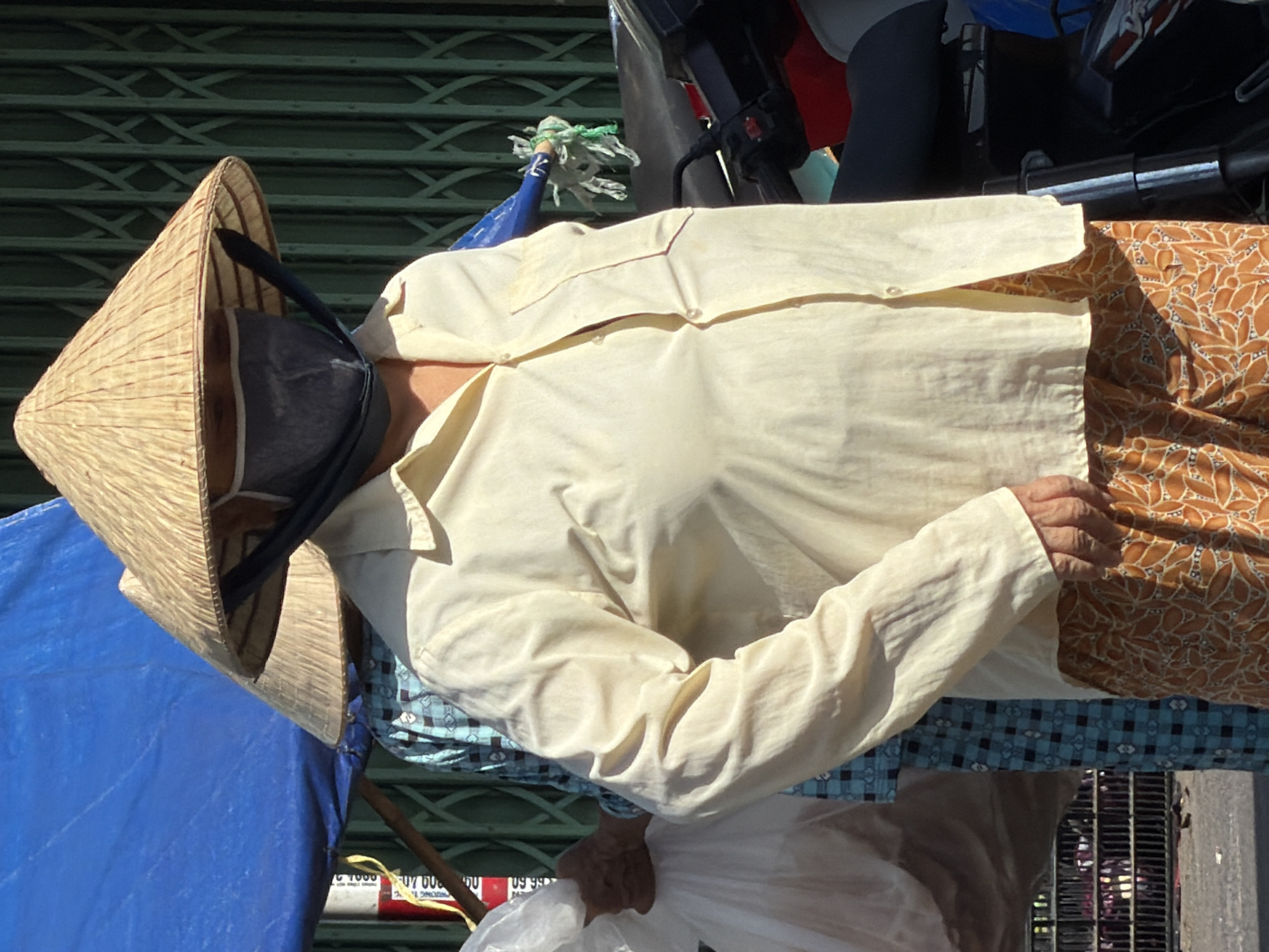 The image depicts a street scene in Nha Trang, Vietnam, likely a market area. In the foreground, a person wearing a traditional conical hat and a light yellow collared shirt is partially visible. The person also wears a dark grey face mask. Their lower body is covered by a patterned orange fabric. To the left of the main subject, another person wearing a blue and white checkered shirt is visible, also wearing a conical hat. This person is holding a transparent plastic bag filled with items. In the background, a green metal gate structure is present, along with parts of a motorcycle. A blue tarp is visible overhead. The lighting suggests it is daytime with sunlight casting shadows.