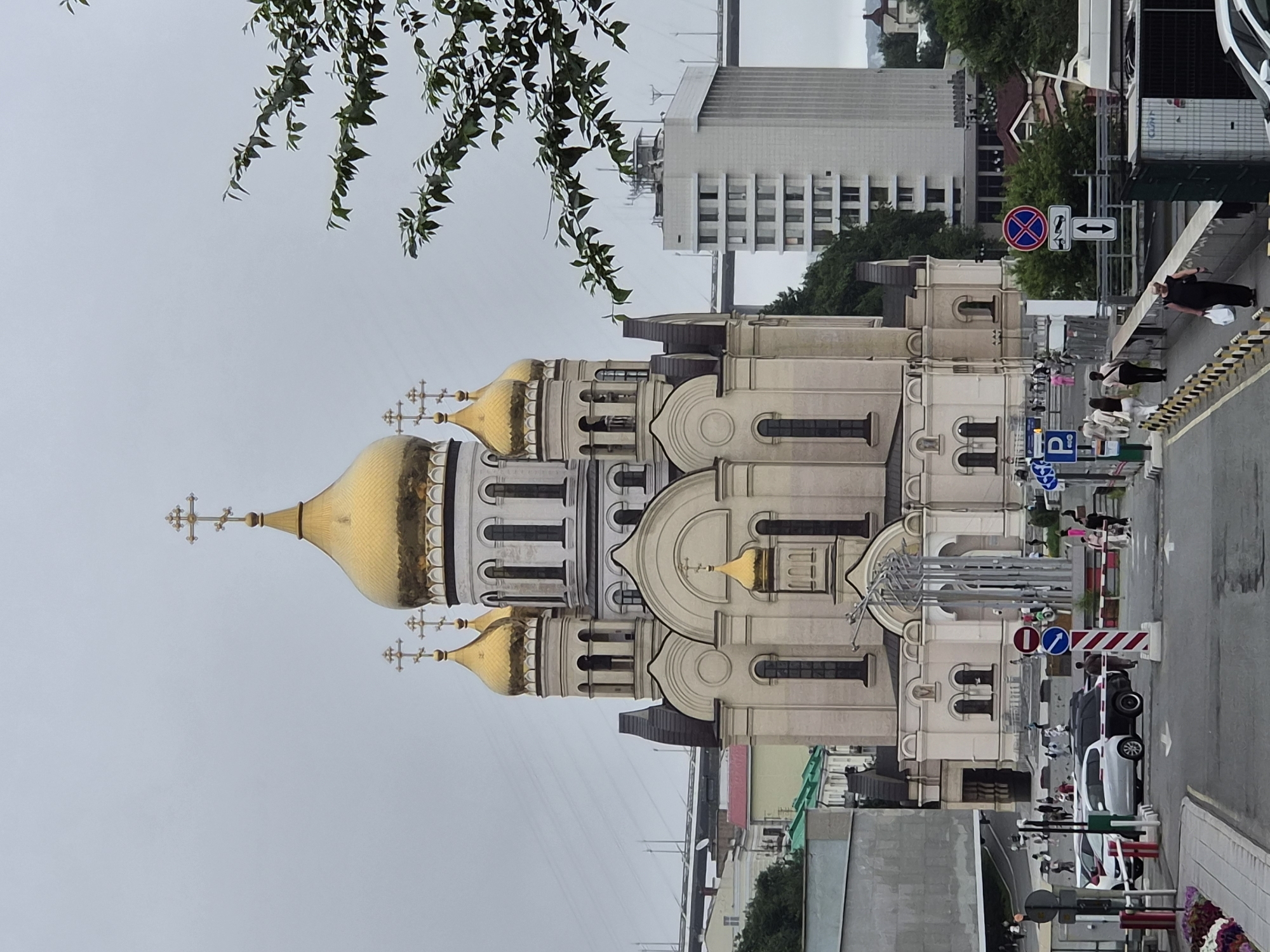 The image depicts the Church of the Transfiguration in Vladivostok, Russia, a prominent landmark characterized by its distinctive golden domes and white facade. Situated on an elevated position, the church overlooks a city landscape that includes modern buildings and a large bridge in the background. The sky is overcast, suggesting a cloudy day.
In the foreground, there is a street with several vehicles, including cars and what appears to be a white SUV. Pedestrians are visible walking along the sidewalk and crossing the street. Traffic signs and barriers are present, indicating a controlled traffic area. One sign depicts a red circle with a diagonal line through a car and a trailer, signifying a prohibition against towing. Another sign shows a blue square with a white letter 'P' for parking, accompanied by an arrow pointing downwards, indicating the direction of the parking area.
The architecture of the church is elaborate, with multiple domes topped by crosses. The main dome is particularly large and golden, standing out against the lighter colored structure of the building. The surrounding buildings are a mix of styles, with some appearing to be residential or commercial, and others having a more institutional look. Green trees and foliage are interspersed throughout the urban environment. The overall scene is a snapshot of daily life in Vladivostok, with the iconic church serving as a central focal point.