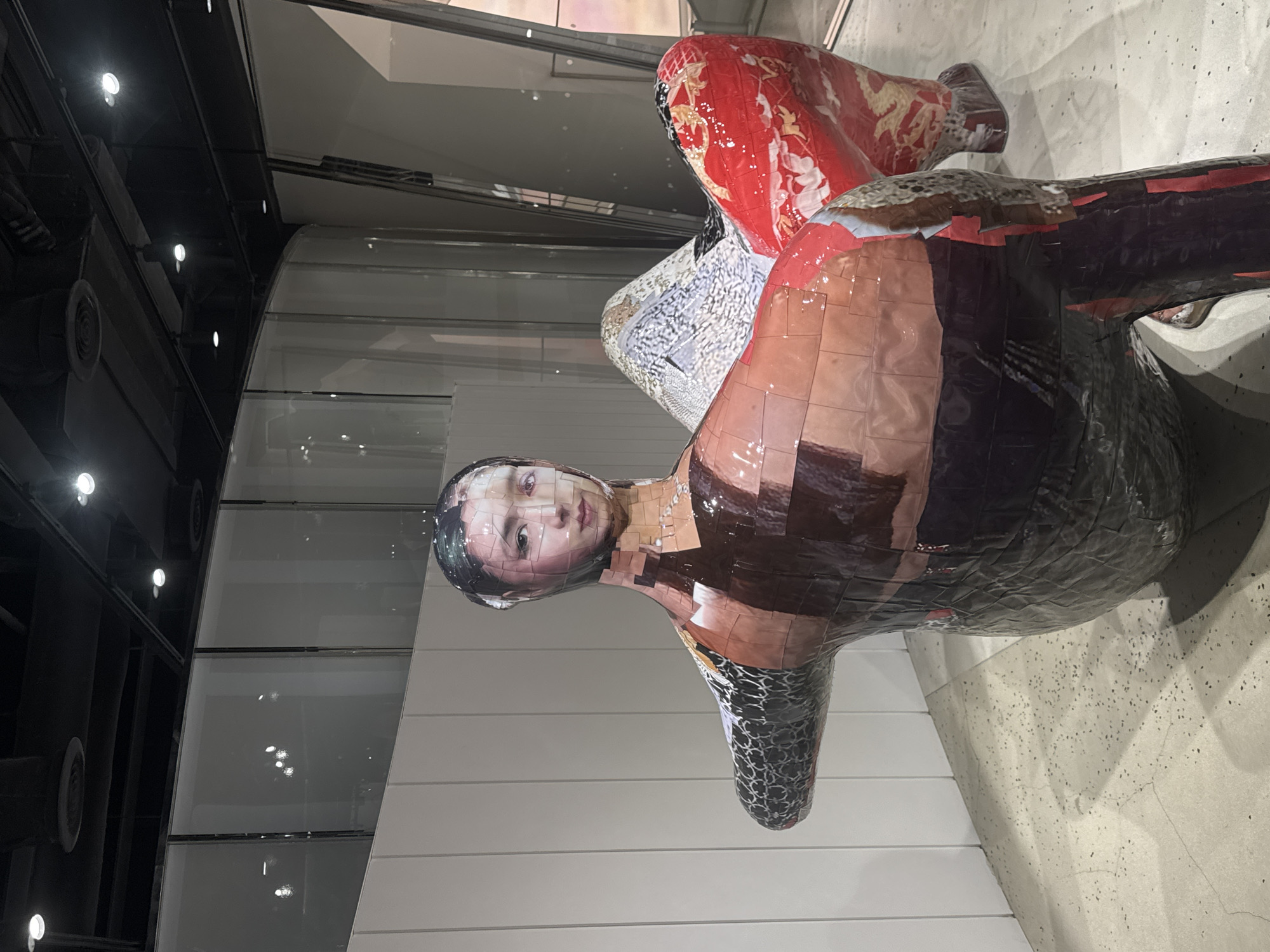 The image shows a sculpture of a human-like figure with a head that appears to be a collage of a woman's face. The body of the sculpture is also made of a collage of various patterns and textures, including what looks like dark scales, brown and red patches, and floral designs. The sculpture is seated or positioned in a way that suggests it is part of a larger installation. The background appears to be an interior space with a white wall on the left, and a curved glass wall or window behind the sculpture, reflecting lights and architectural elements. The floor is a speckled concrete. Based on the context of similar artworks and interior design styles, this sculpture is likely located in the Xintiandi district of Shanghai, China, known for its contemporary art galleries and trendy commercial spaces.