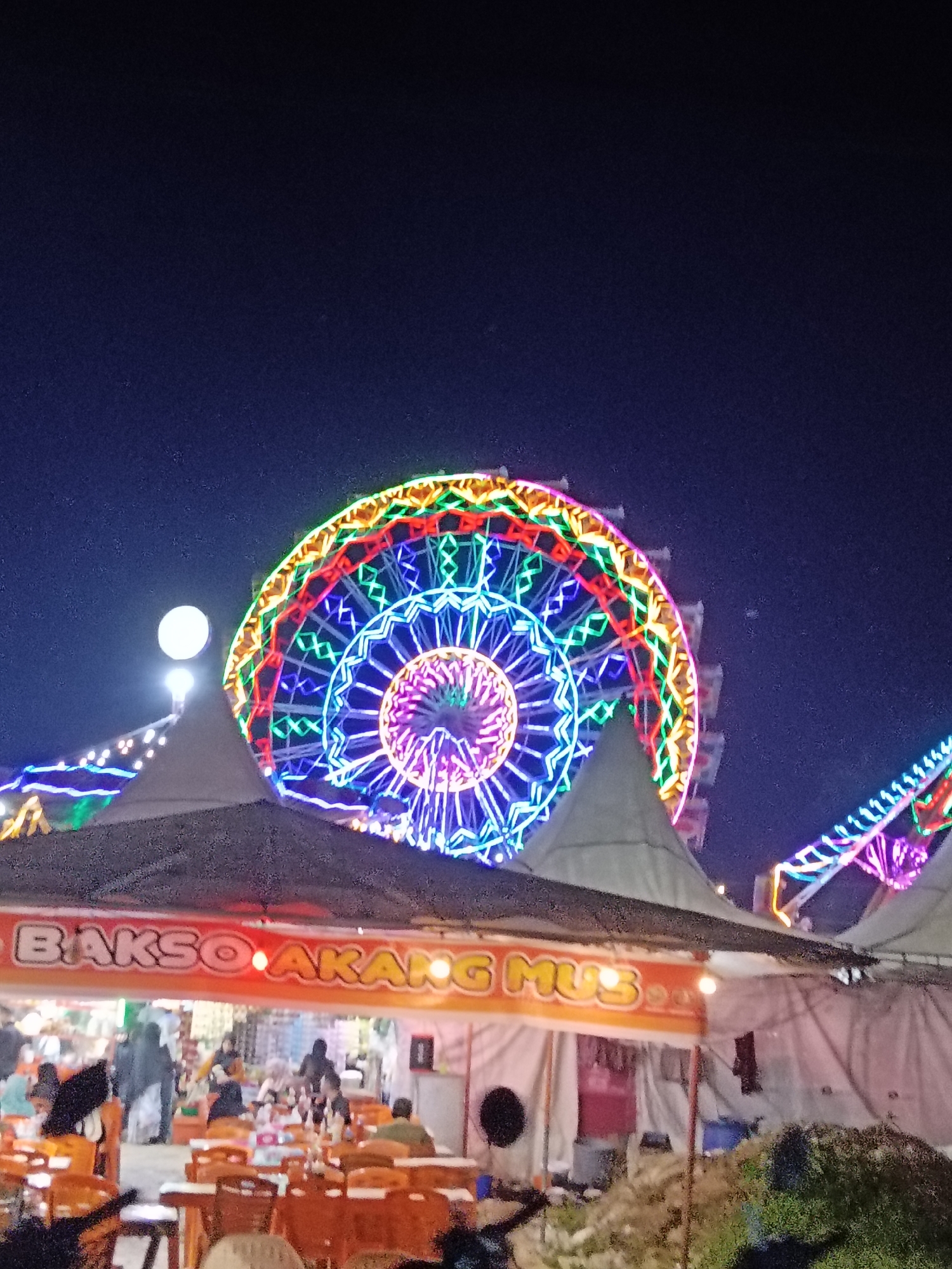 A nighttime scene depicts a brightly lit Ferris wheel with multiple colored neon lights in Keudepanteubreuh, Indonesia. In the foreground, several white tents with dark patterned roofs are set up, possibly for food stalls. One tent displays an orange banner with text that reads "Bakso Akar Mus." Underneath the tents, tables and chairs are visible, with some people seated and others standing. Further in the background, more tents and a section of a ride with neon lights can be seen. The sky is dark blue, indicating it is nighttime.