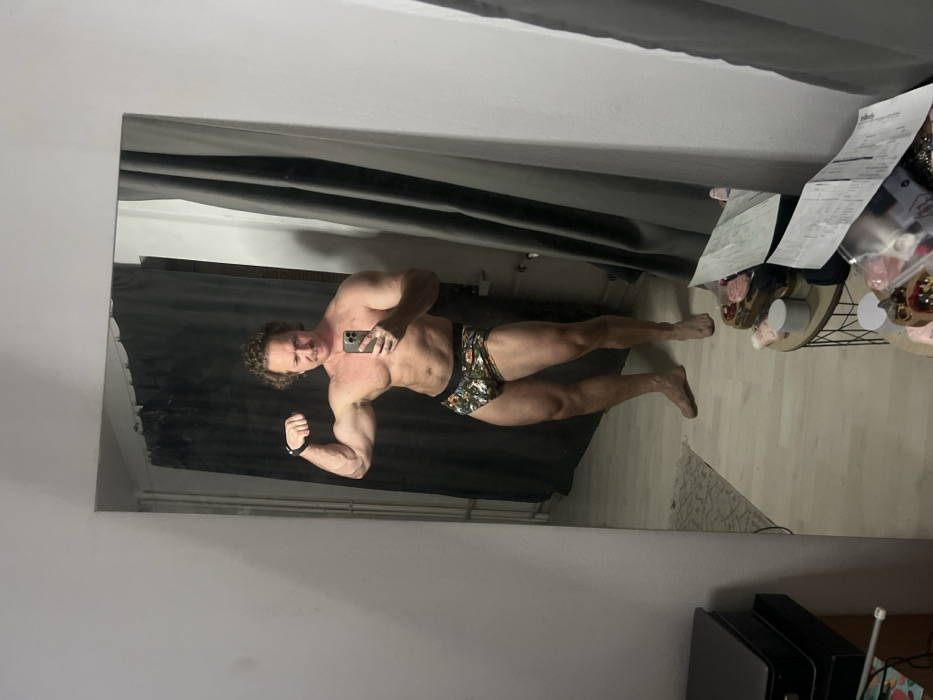A muscular man poses in a mirror. He flexes his right arm, holding a phone in his left hand. He wears patterned boxer briefs. The background includes a curtain, a small table with items, a bill, and a portion of flooring. The mirror's reflection shows parts of the room including walls and a dark-colored object. The image is slightly tilted.