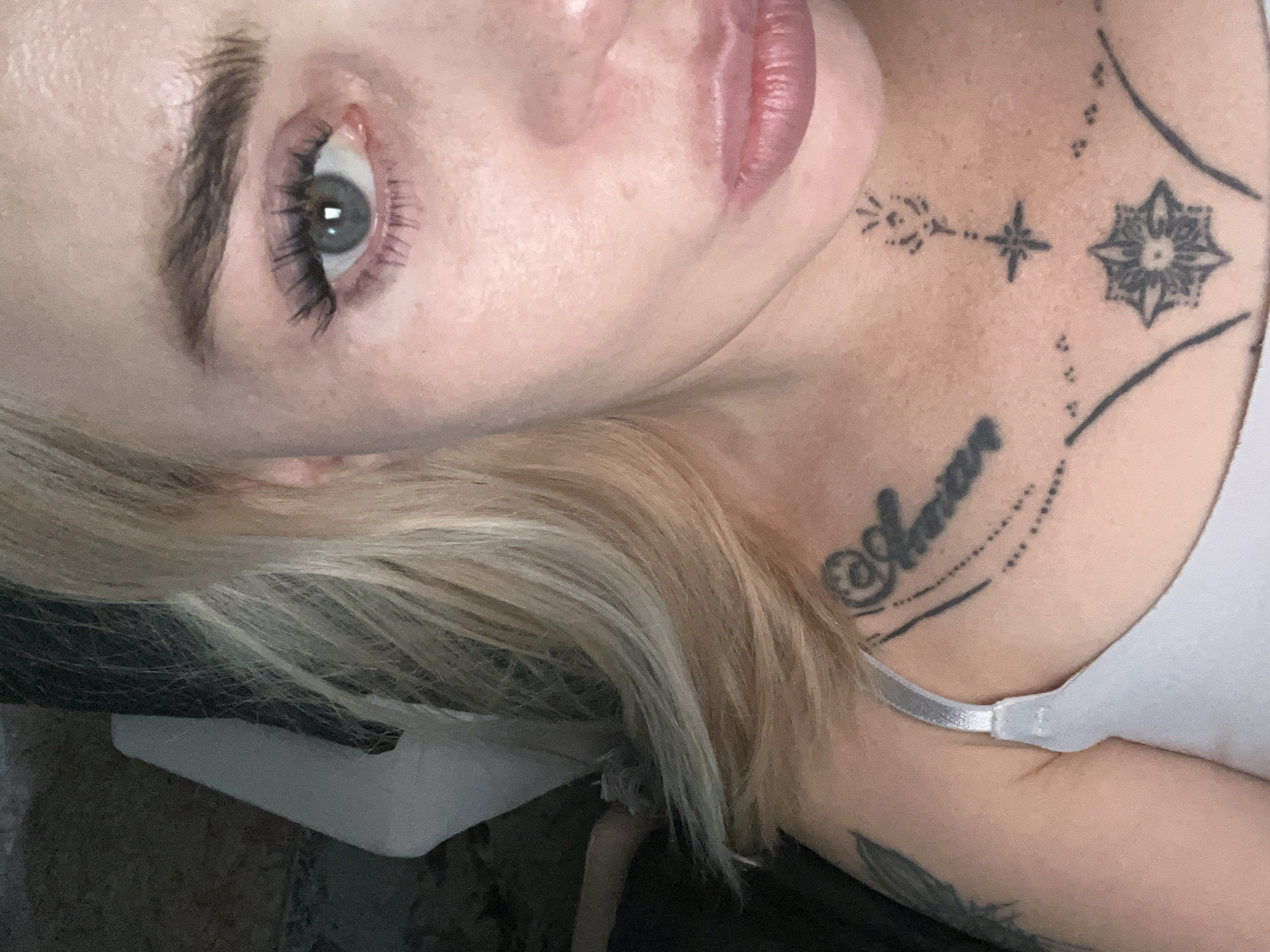 A medium close-up, high-angle shot captures the right side of a fair-skinned woman's face, from her eye to her collarbone. Her blonde hair, parted on the left, frames her face and falls over her shoulder. Her right eye, wide open and looking towards the camera, is a striking blue-grey. Her eyebrows are dark and well-defined, and she has long, dark eyelashes. Her skin is fair with some freckles visible. Her lips are full and painted with a muted pink lipstick. A thin, silver bra strap is visible just below her collarbone. She has a tattoo of the word "Amore" in cursive script on her upper chest, above a star design and a floral mandala tattoo. To the left of the frame, a dark, textured surface with a white object is partially visible, suggesting an indoor setting.