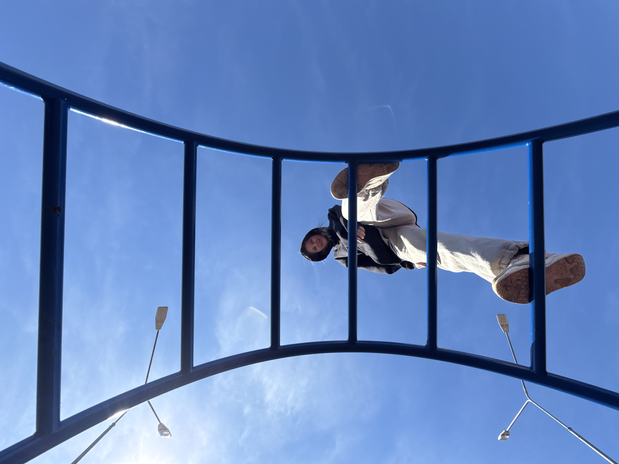 A low-angle shot looking up shows a person on a blue metal climbing structure against a bright blue sky. The person is wearing light-colored pants and sneakers, with a dark jacket or hoodie. Two lights and their support arms are visible through the structure, backlit by the sun. The city of Горный, Russia is not visually discernible.