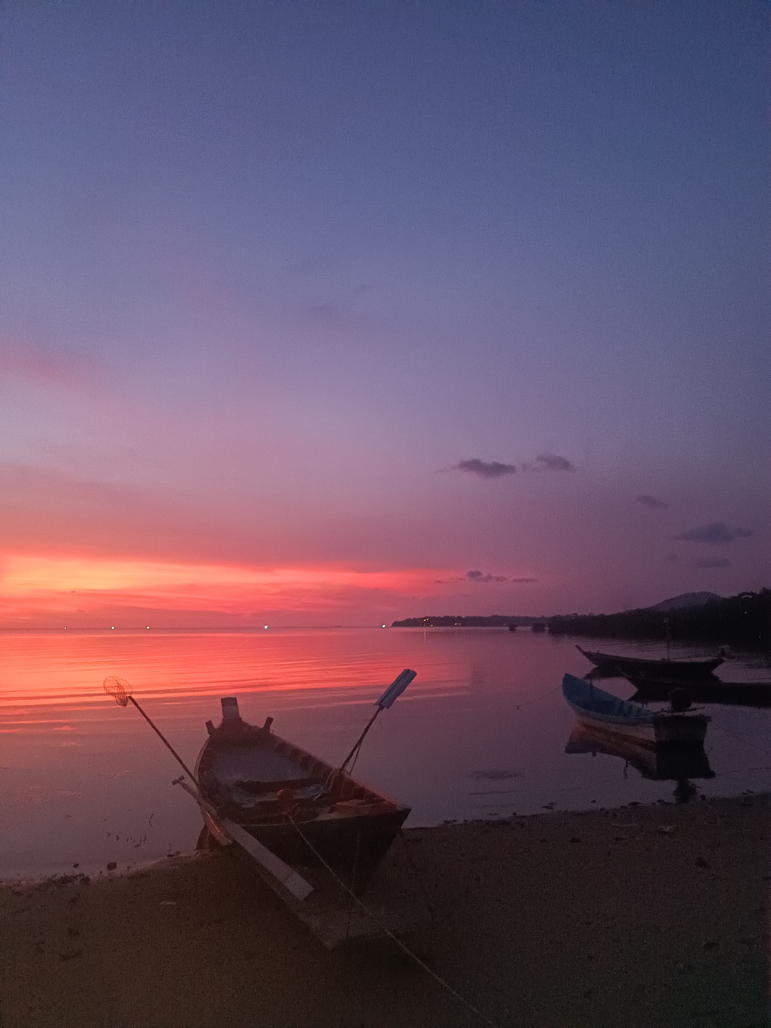 The image depicts a coastal scene at dusk in Baan Wok Tum, Thailand. The sky transitions from a deep blue/purple to a vibrant orange/red at the horizon, reflecting onto the calm water. In the foreground, a small wooden boat rests on the sandy shore, with oars or poles extending upwards. Further out, two more boats are seen floating on the water. The shoreline curves towards the right, with trees and possibly a small settlement visible in the distance. The lighting suggests either sunrise or sunset, emphasizing the serene atmosphere.