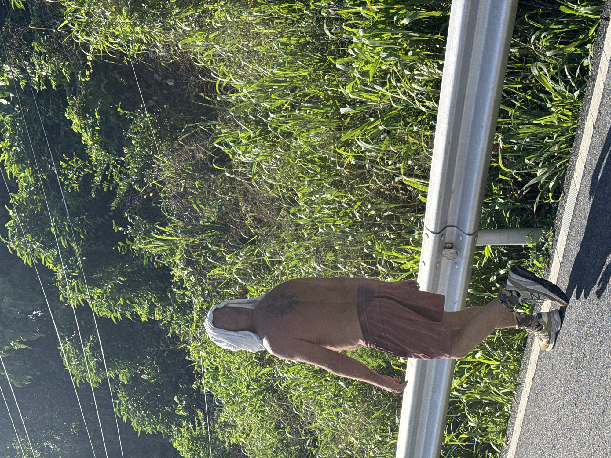 A shirtless man with a white towel draped over his head and shoulders walks along a road in Rawai, Thailand. He has a large, dark tattoo on his back. He is wearing red shorts and athletic shoes. The man is walking away from the camera on the right side of the road, next to a metal guardrail. Dense green foliage lines the side of the road behind the guardrail, and power lines are visible in the background against a bright sky. The sun is casting shadows on the road.