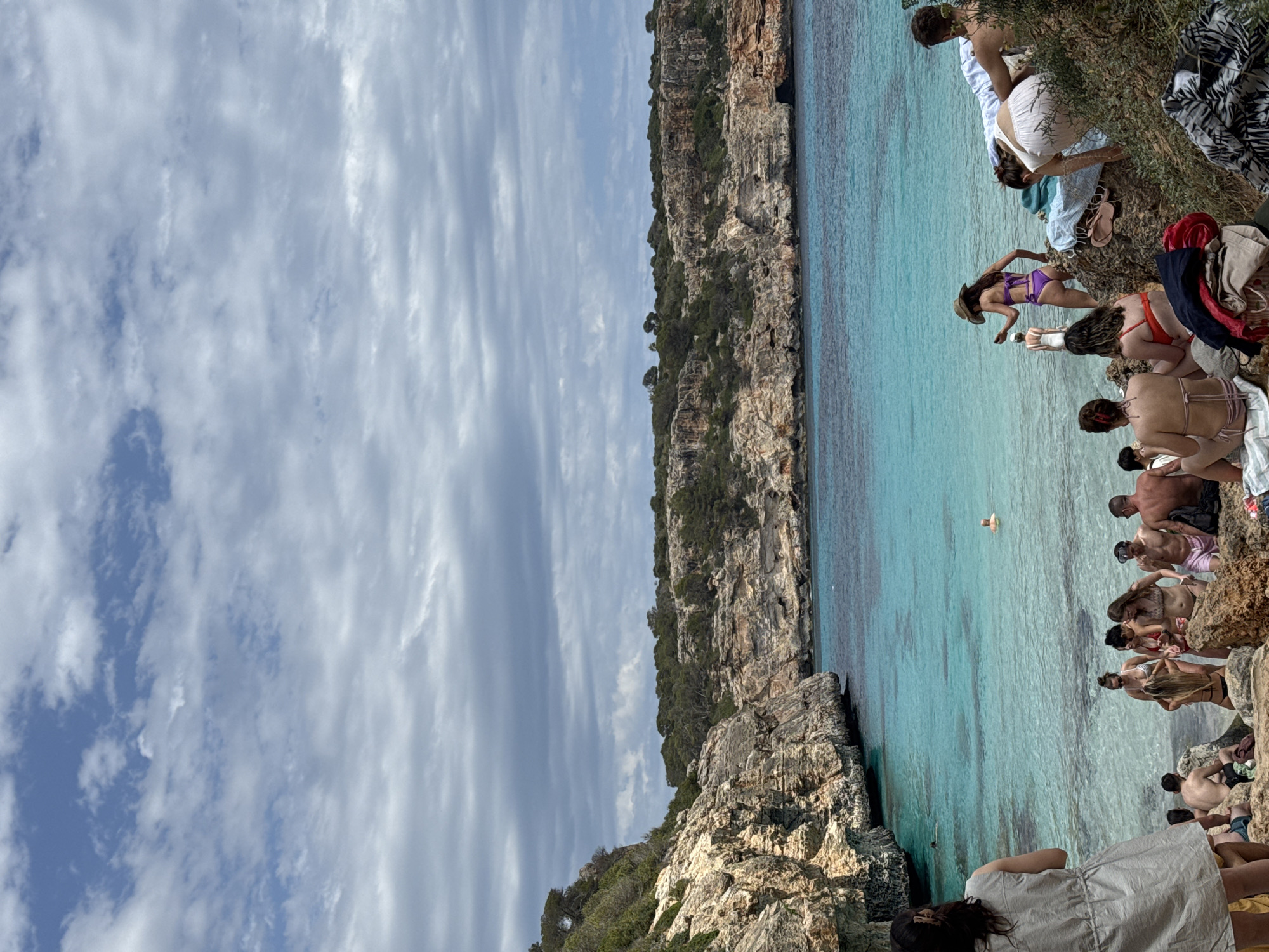 The image captures a coastal scene, possibly a cove or bay, within Santanyí, Spain. Clear, turquoise water fills the frame, meeting rocky cliffs on both sides. Several people are positioned along the cliff edges, some appearing to be entering or exiting the water, while others are sunbathing or socializing on the rocks. The sky is partially cloudy, with the cloud formations dominating the left side of the image. The overall atmosphere suggests a popular beach or swimming spot during a warm day.