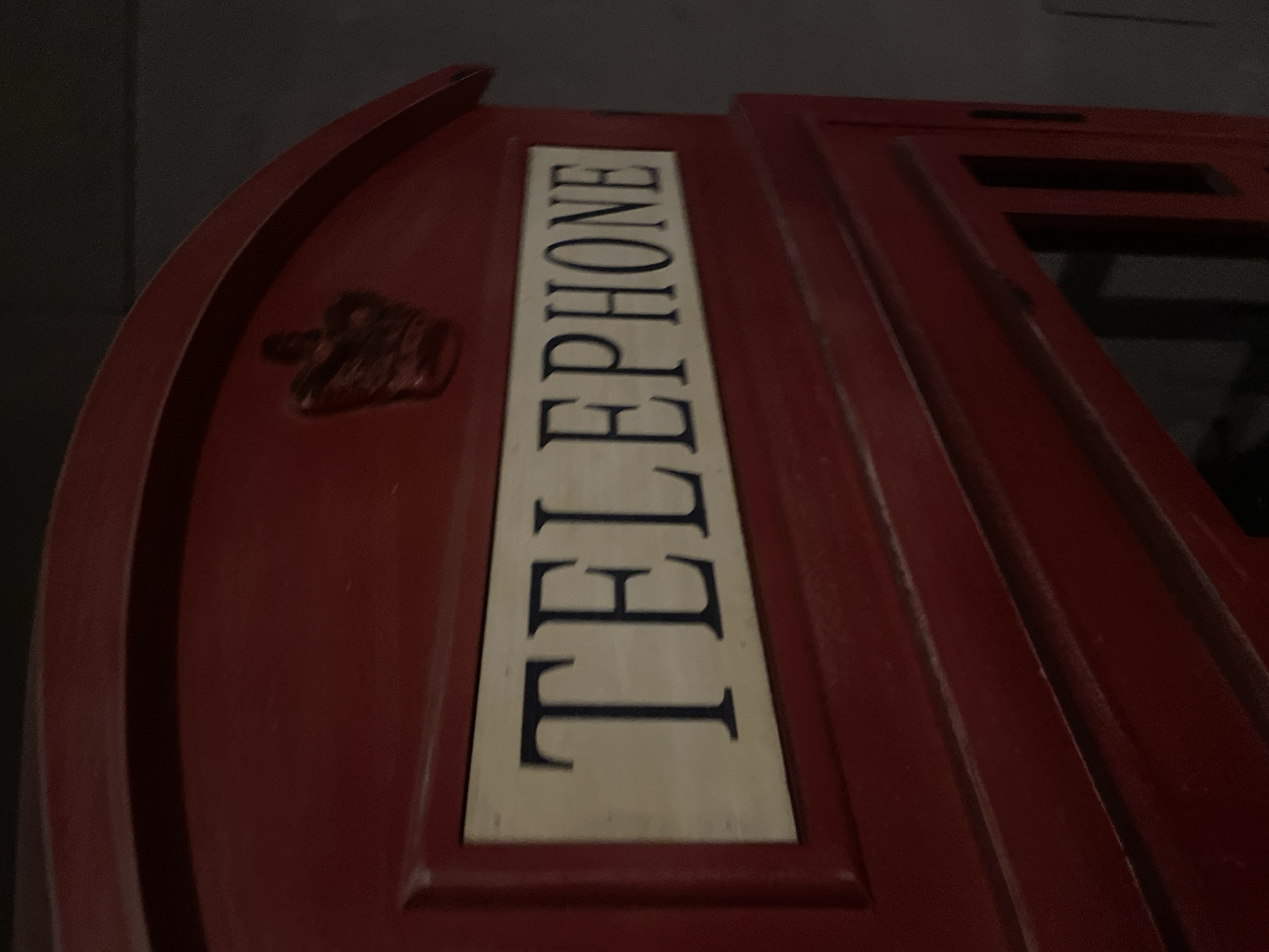 The image shows a close-up of a red telephone booth, specifically focusing on the upper portion. The word "TELEPHONE" is vertically oriented on a white rectangular panel. To the left is a raised emblem of a crown. The surface has multiple vertical ridges.
It is not possible to determine the location of this telephone box being in the city of Puebla, Mexico.