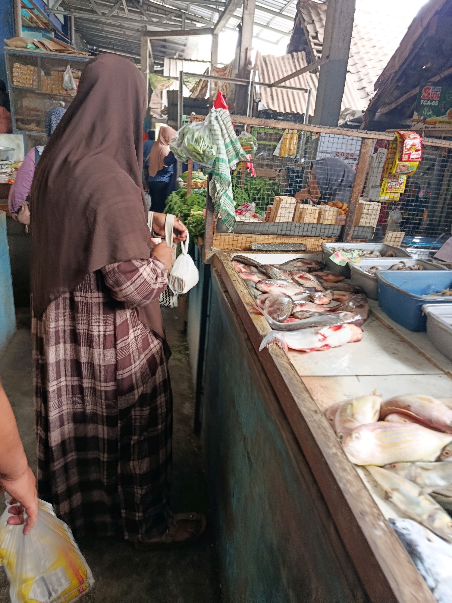 The image depicts a market stall in Bululawang, Indonesia, with a focus on fresh produce and seafood. A person wearing a brown headscarf and a plaid dress stands on the left, holding white plastic bags. To their right, a display of various fish is laid out on a counter, some in plastic containers. Behind the fish, more produce, including leafy greens, is visible. The market appears to be an open-air structure with a corrugated roof and wooden stalls. Other individuals are present in the background, engaged in market activities. The scene captures a moment of commerce and daily life in the Indonesian market.