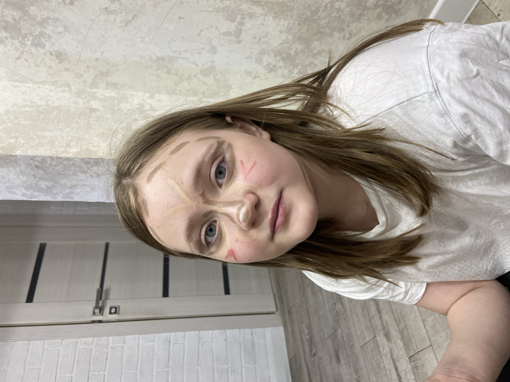 A young woman with light skin and brown hair is sitting indoors. She is wearing a white t-shirt. The woman has makeup applied to her face in a style that appears to be for contouring, with light and dark shades of makeup visible on her forehead, nose, cheeks, and around her eyes. The background includes a door with dark accents and a tiled wall. The overall location appears to be a residential setting. It is not possible to confirm if the scene is located in Chaikovsky, Russia, without further information.