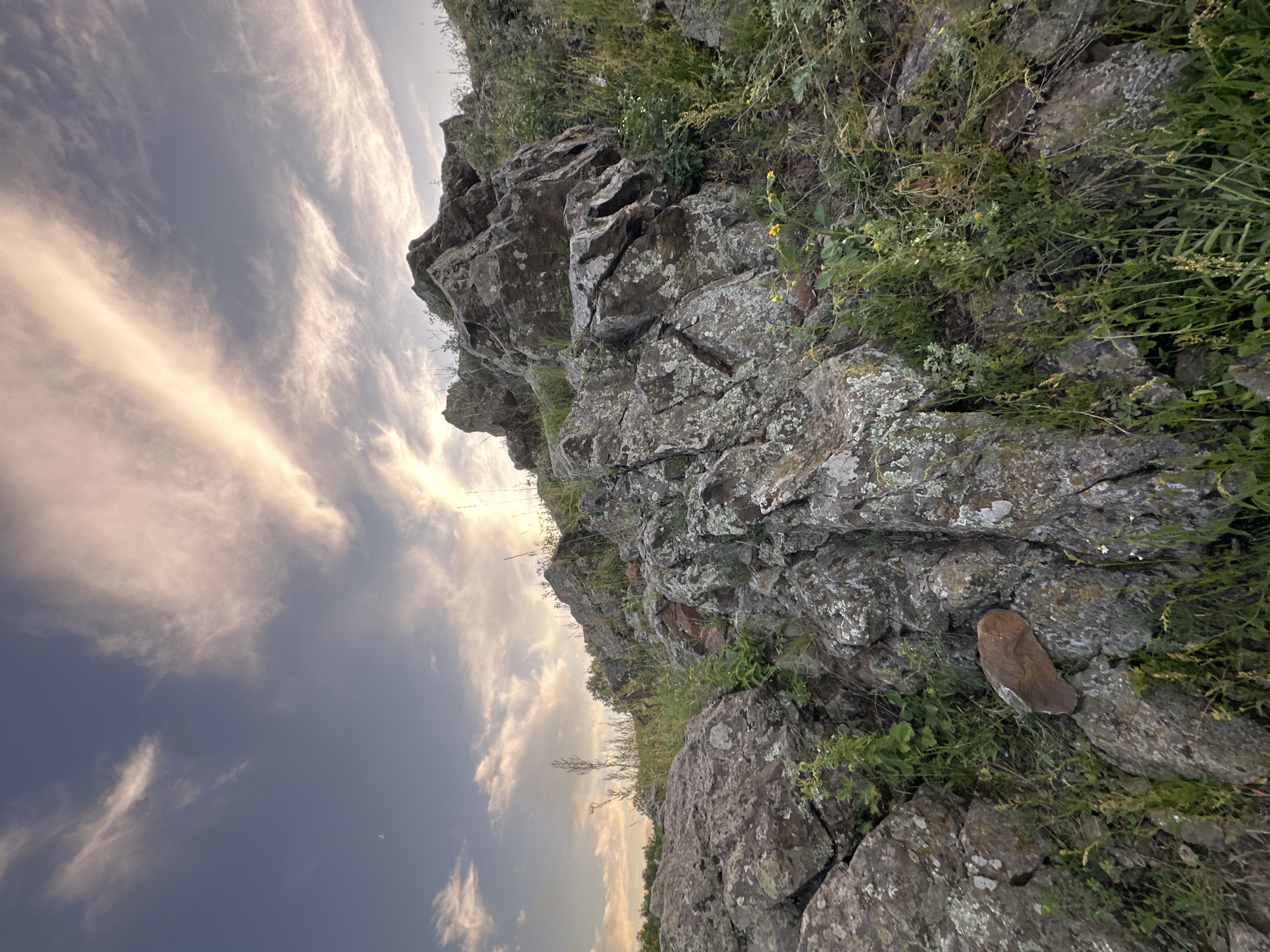 The image depicts a rocky landscape with vegetation under a sky with wispy clouds. Large, weathered rocks with patches of lichen dominate the foreground and midground, interspersed with green grass and some small yellow wildflowers. In the distance, more rocks and trees can be seen. The sky above is a gradient of blue and pale orange, with thin, elongated clouds catching the light. A sliver of the moon is visible in the upper left quadrant of the sky. The lighting suggests it may be near sunset or sunrise. There are no people or man-made structures visible in the image.