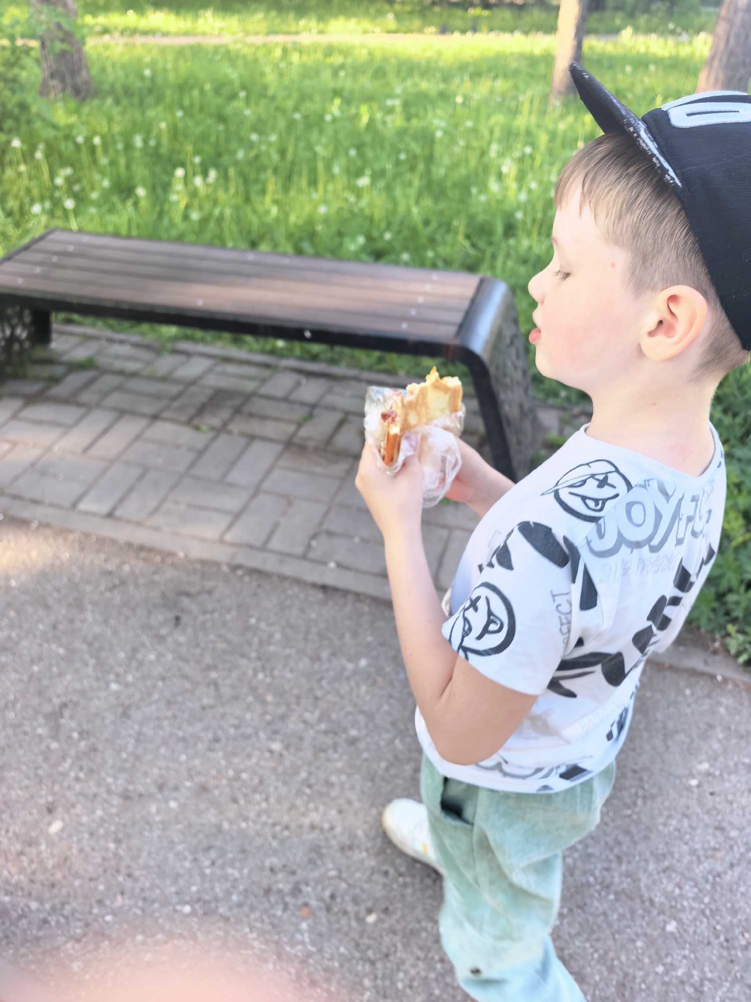 The image shows a young boy standing in a park in Sterlitamak, Russia. He is wearing a white t-shirt with a cartoon print, light green pants, and white shoes. He has a black cap on his head. The boy is holding a sandwich wrapped in plastic in his hands. Behind him, there is a wooden bench on a paved pathway, with green grass and trees in the background. The scene appears to be outdoors on a sunny day.
