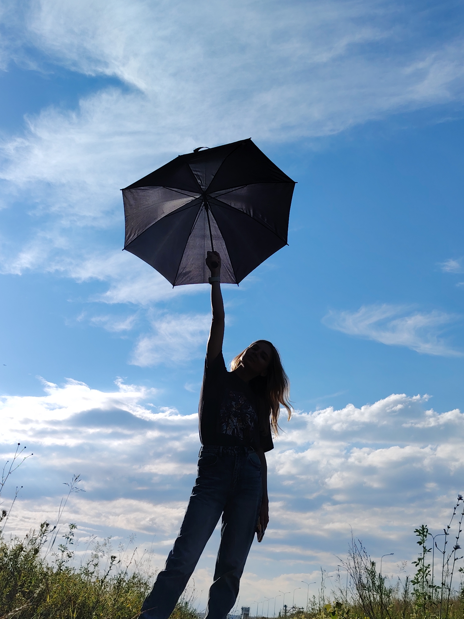 A person is silhouetted against a bright blue sky with scattered white clouds, holding a black umbrella above their head with their right hand raised. The person is wearing a dark t-shirt and blue jeans. The lower portion of the image shows a grassy field with some dry vegetation and wildflowers. In the background, beyond the field, is a road with streetlights and a distant structure that appears to be a bridge or overpass. The image is taken from a low angle, looking up at the person and the sky. The location appears to be an outdoor, open area near a road, possibly on the outskirts of Chaikovsky, Russia, given the presence of streetlights and infrastructure commonly found in urban or suburban environments.