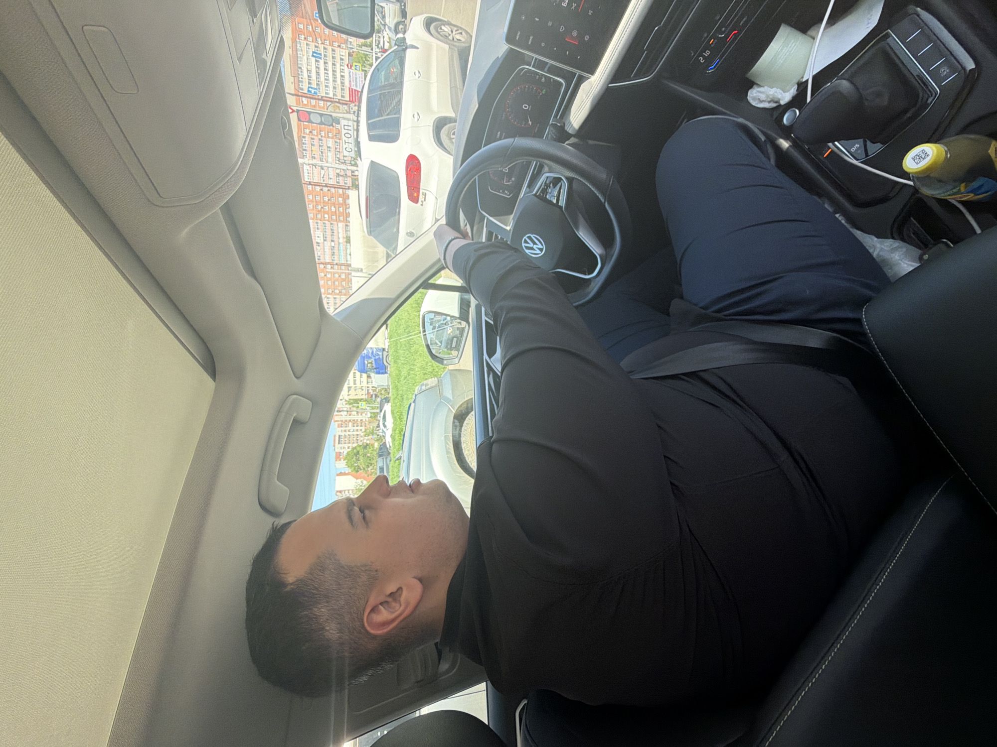 A man is seated in the driver's seat of a car, facing forward and slightly to his left, looking out the window. He is wearing a dark long-sleeved shirt and dark trousers, with a seatbelt fastened across his chest. His right hand rests on the steering wheel, which bears a Volkswagen logo.
The car's interior is visible, featuring a digital dashboard, a gear shifter, and a climate control panel displaying "21.0". On the center console, a yellow-capped bottle with a QR code and a roll of tissue are present. The seats are black leather.
Outside the car, other vehicles are visible, suggesting a street or parking area. Multi-story buildings line the background. The scene takes place during bright daylight, indicated by strong sunlight and shadows. The image has a rotated orientation.
A red sign outside the car clearly displays the Cyrillic word "СТОП," meaning "STOP."