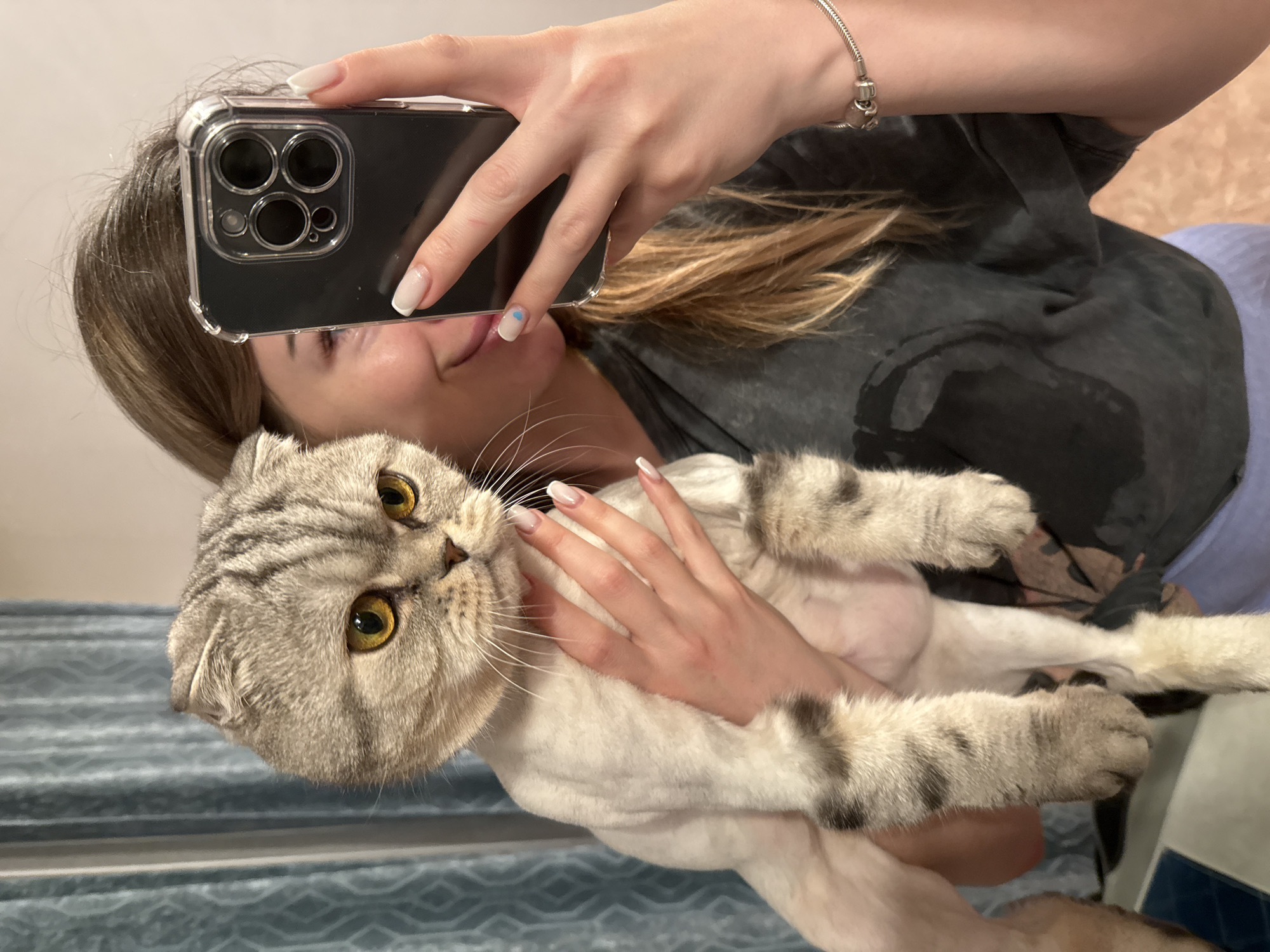 A person is taking a selfie with a gray and white tabby cat. The person is holding a phone with multiple camera lenses. The person has long blonde hair and manicured nails. The cat has yellow eyes and a gray tabby coat. The person is wearing a gray shirt. The background is a light blue wall with horizontal stripes. The location is unknown.