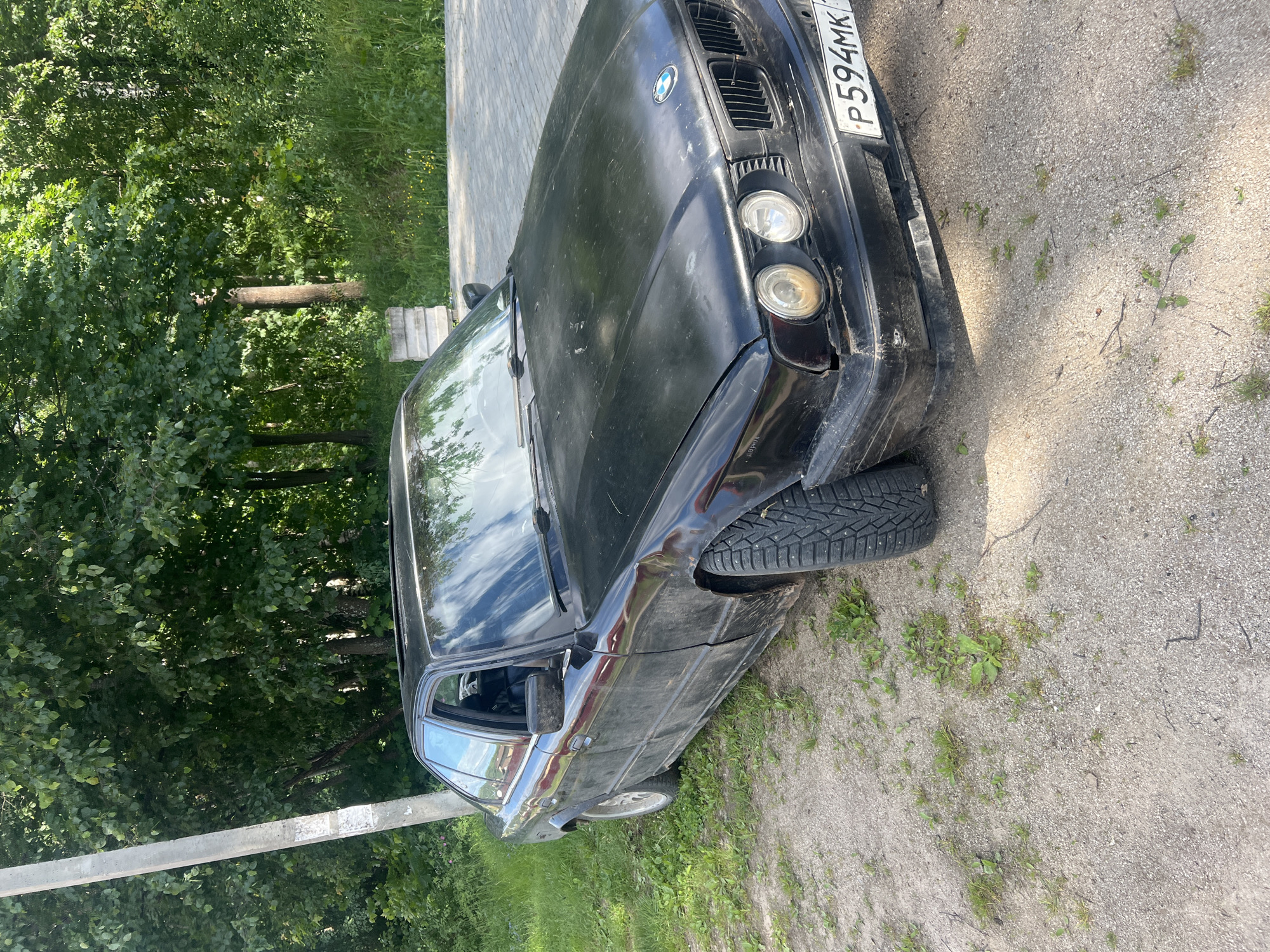 The image displays a black, visibly dirty and worn BMW E34 sedan parked outdoors on a sunny day in the city of Noviy, Russia. The vehicle is situated on a surface composed of light-colored gravel and dirt, interspersed with patches of green grass and weeds. To the right of the car, a paved area constructed with gray interlocking pavers is visible.
The BMW's front features a blacked-out kidney grille, two sets of dual round headlights (the left outer headlight appears yellowed), and a Russian license plate displaying 'P594MK 117'. The car's body shows a faded, matte black finish and accumulated dirt. The front right tire has a pronounced, aggressive studded winter tread pattern. A sunroof is visible on the vehicle's roof.
In the background, a dense line of lush green trees forms a natural barrier, suggesting a wooded or heavily vegetated environment. On the far left, a tall, light-colored concrete utility pole stands vertically. A smaller, weathered concrete block structure is partially visible near the paved area on the right, behind the car's windshield. No individuals are present in the scene.