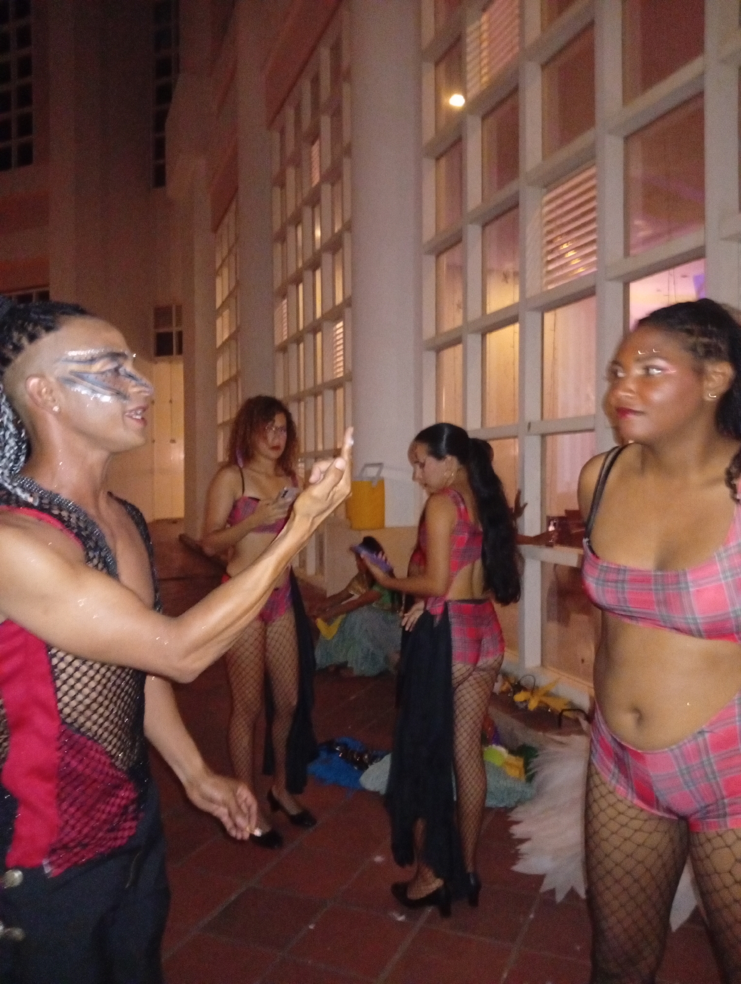 The image shows a group of four individuals in a room with a tiled floor and a large wall of windows. The individuals are wearing costumes or attire reminiscent of Carnival or a similar festive event. The person closest to the camera is wearing a red and black mesh top and has elaborate makeup. Three other individuals, who are wearing matching plaid red tops and shorts and fishnet stockings are also present. The image appears to have been taken at night or in a dimly lit environment, with the windows showing external lighting. The location is in the city of Pedro González, Venezuela.