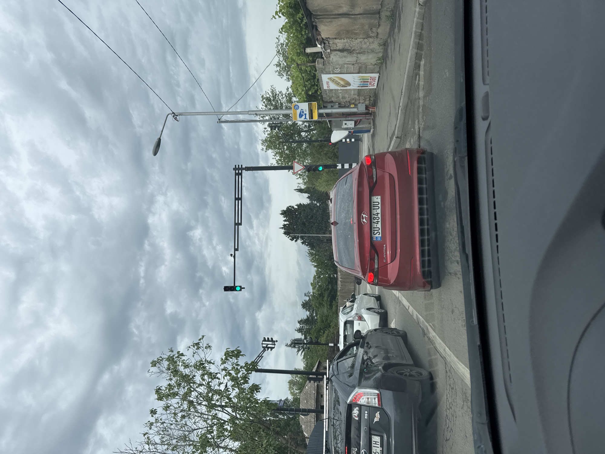 The image shows a traffic jam on a cloudy day in an urban environment. In the foreground, the dashboard of a car is visible. Ahead of this car, several vehicles are stopped in traffic, including a red Hyundai Elantra, a dark gray Toyota Prius, and a white car. Traffic lights are visible overhead, with a green light illuminated on one signal. Trees line the side of the road, and buildings, including a wall with advertisements, are visible in the background. The street is paved, and there is a sidewalk. The overall impression is of a typical city street scene during a period of congestion.