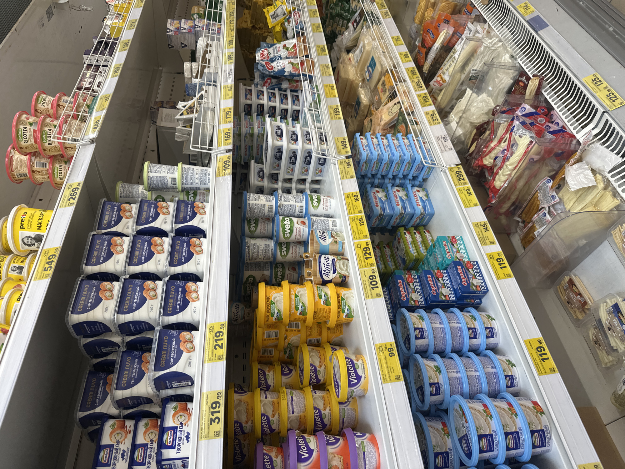 The image displays a well-stocked supermarket shelf in Novoaltaysk, Russia, showcasing a variety of dairy products, primarily cheese and cream cheese. The shelves are organized with numerous products neatly arranged in their packaging. Prominent brands visible include "Cream Nuvo," "Hochland," "Violette," and "Bonfesto Ricotta." Prices are displayed on yellow tags, indicating a commercial setting. The lighting suggests an indoor environment, typical of a grocery store. No people or interactions are visible, and the scene is static. The products are predominantly packaged in plastic containers and tubs, with some in resealable bags. The overall impression is one of a commercial food retail environment.