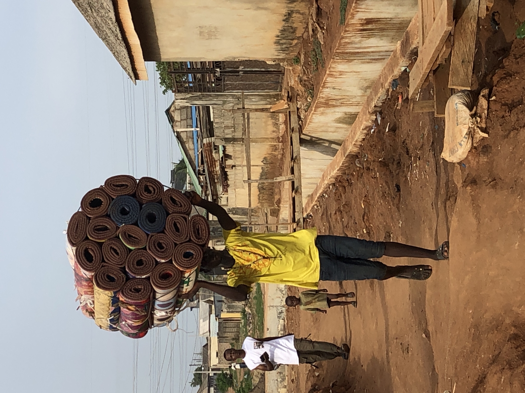 A man in a yellow shirt carries a large bundle of rolled mats on his head down a dirt road in a city of Unknown, Unknown. The bundle is roughly spherical, made of many brown and blue rolled mats. The man wears blue jeans and sandals. Another man in a white shirt stands on the road, and a child in a green shirt stands further down. Buildings line the road, including some made of corrugated metal. A cement bag sits on the side of the road. Overhead power lines are visible.