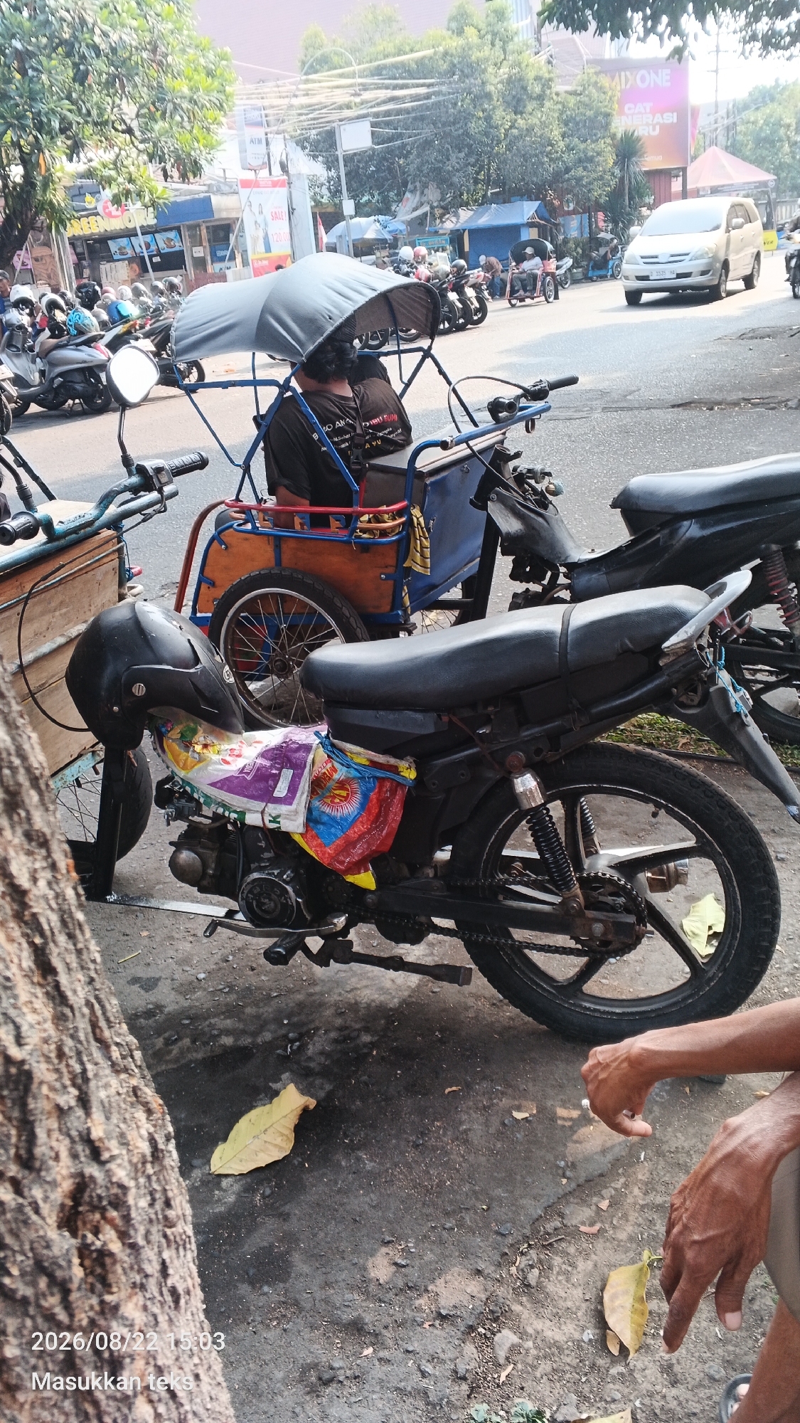 The image shows a street scene in Malang, Indonesia, with a focus on a tricycle-like vehicle (known locally as a "becak") parked next to a motorcycle. The becak has a dark-colored canopy and appears to be waiting for passengers. A person is seated in the becak, wearing a dark t-shirt. A black helmet is placed on the handlebars of the motorcycle, which is decorated with a colorful plastic bag. Other motorcycles and vehicles are visible in the background, along with some buildings and trees, suggesting a typical urban environment. The ground is paved with asphalt and shows signs of wear. Fallen leaves are scattered on the ground. The date stamp in the lower left corner indicates "2026/08/22 15:03".